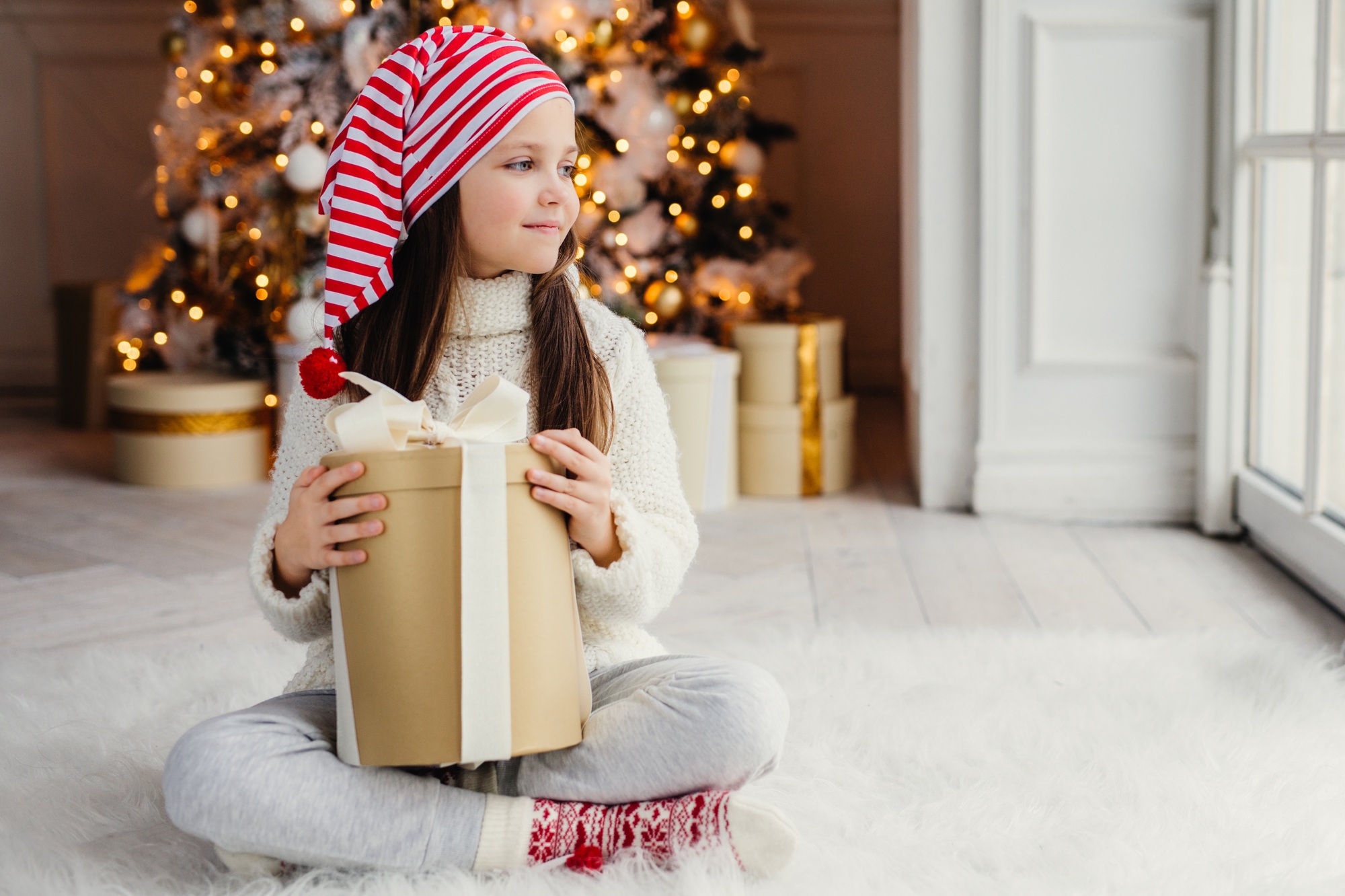 Adorable female child wears santa claus hat, warm clothes, hold wrapped gift box