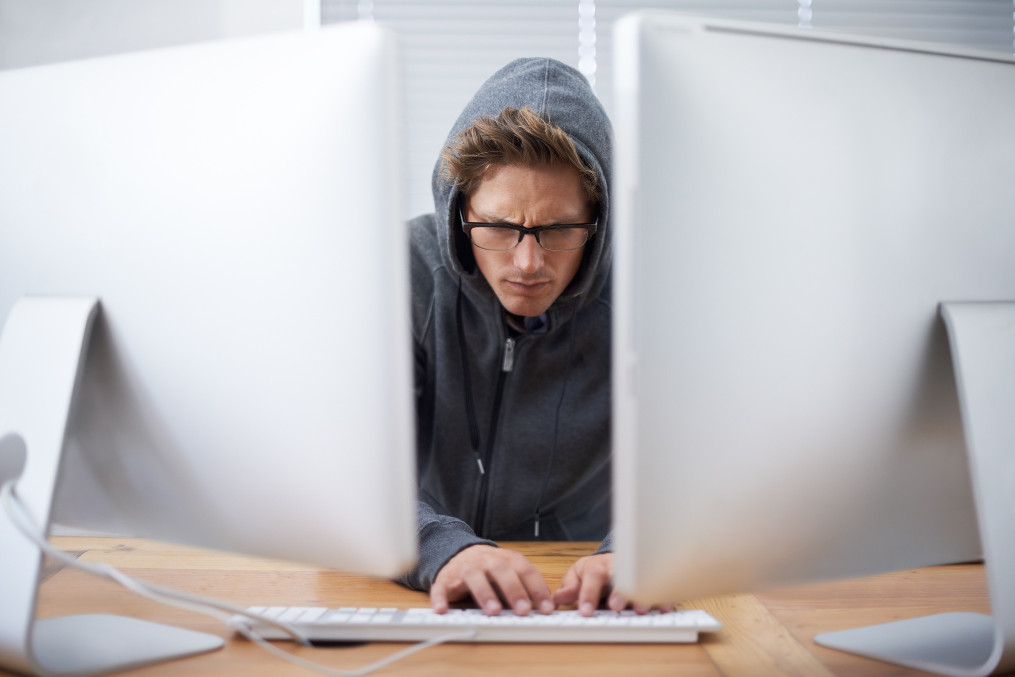 Burning the midnight oil. A young hacker working on dual screens and a laptop wearing a hoodie.