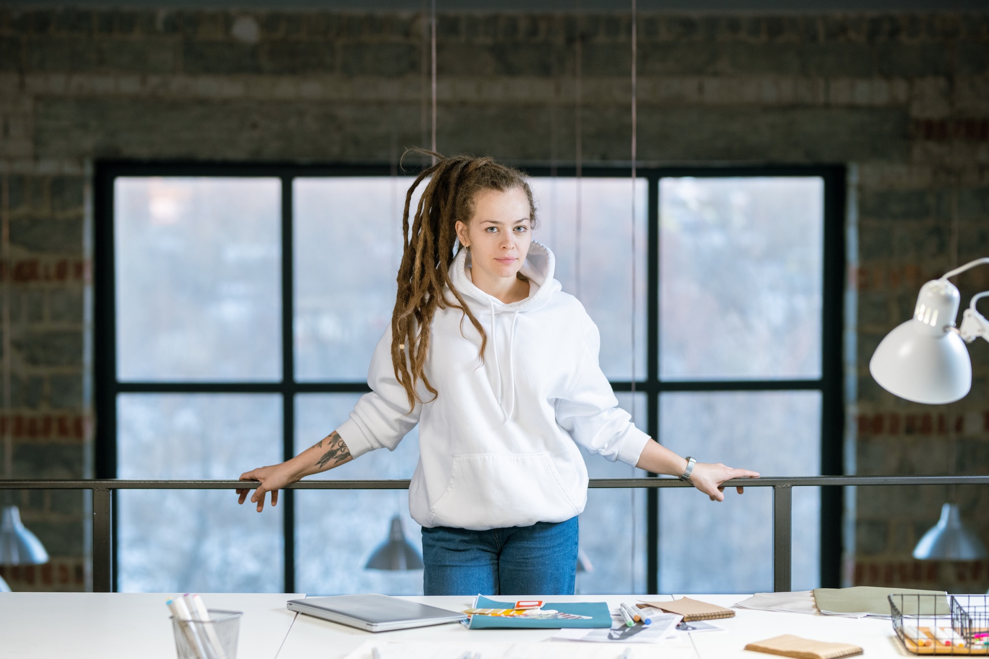 Pretty young creative designer in white hoodie and blue jeans standing in studio