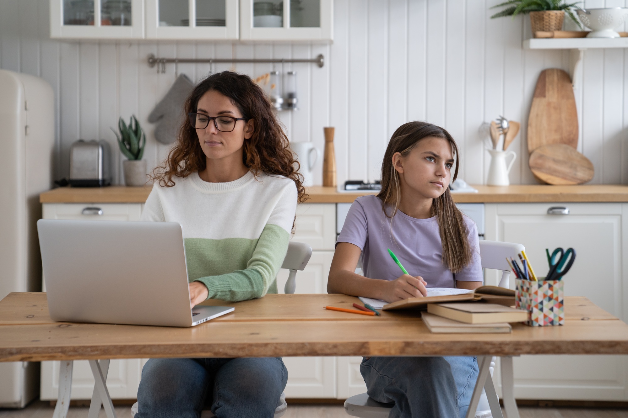 Successful focused business woman mom doing work at laptop sits at kitchen table near with daughter