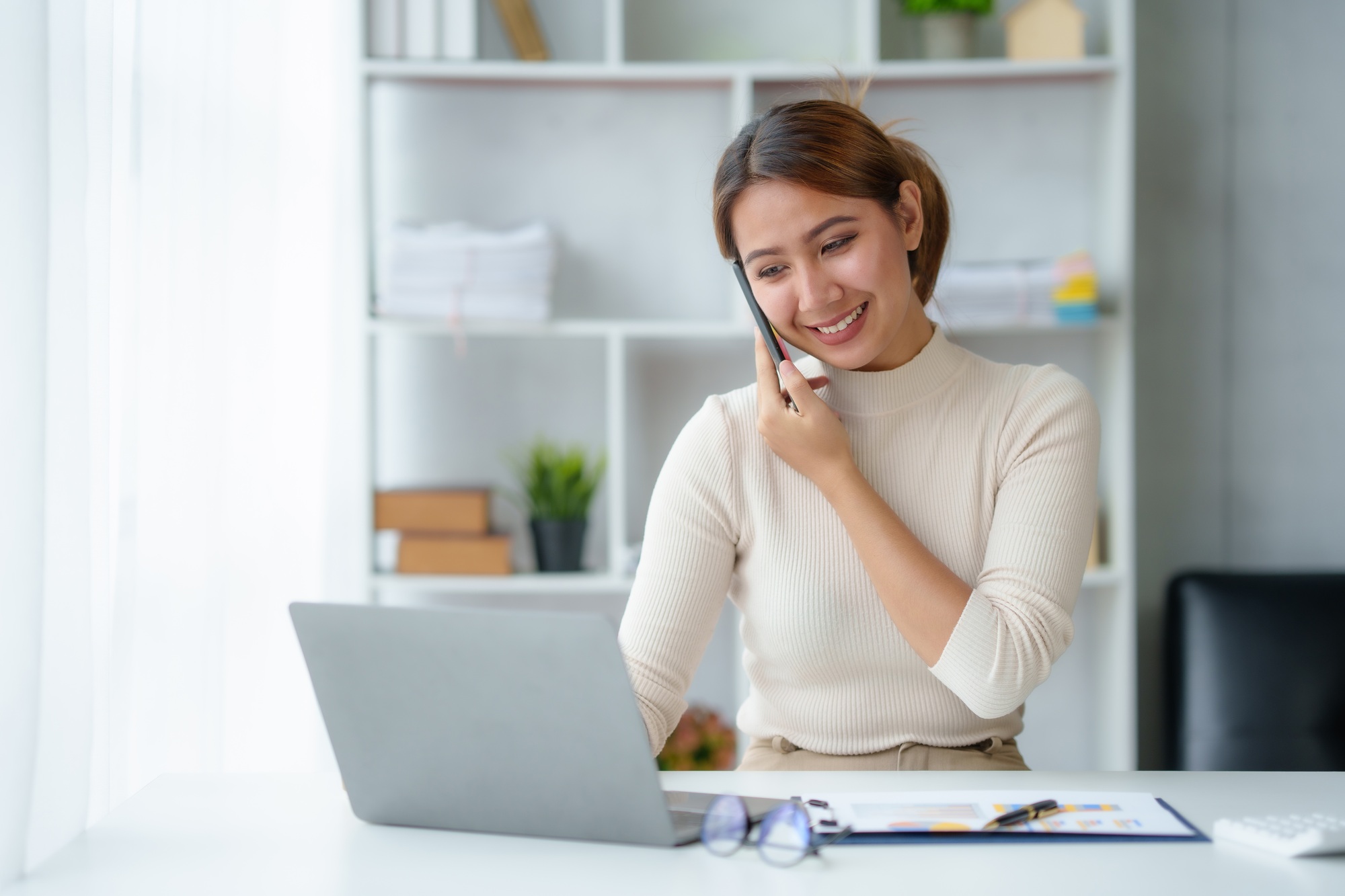 Young Asian businesswoman sit with her laptop computer happily talking on the phone with her custome