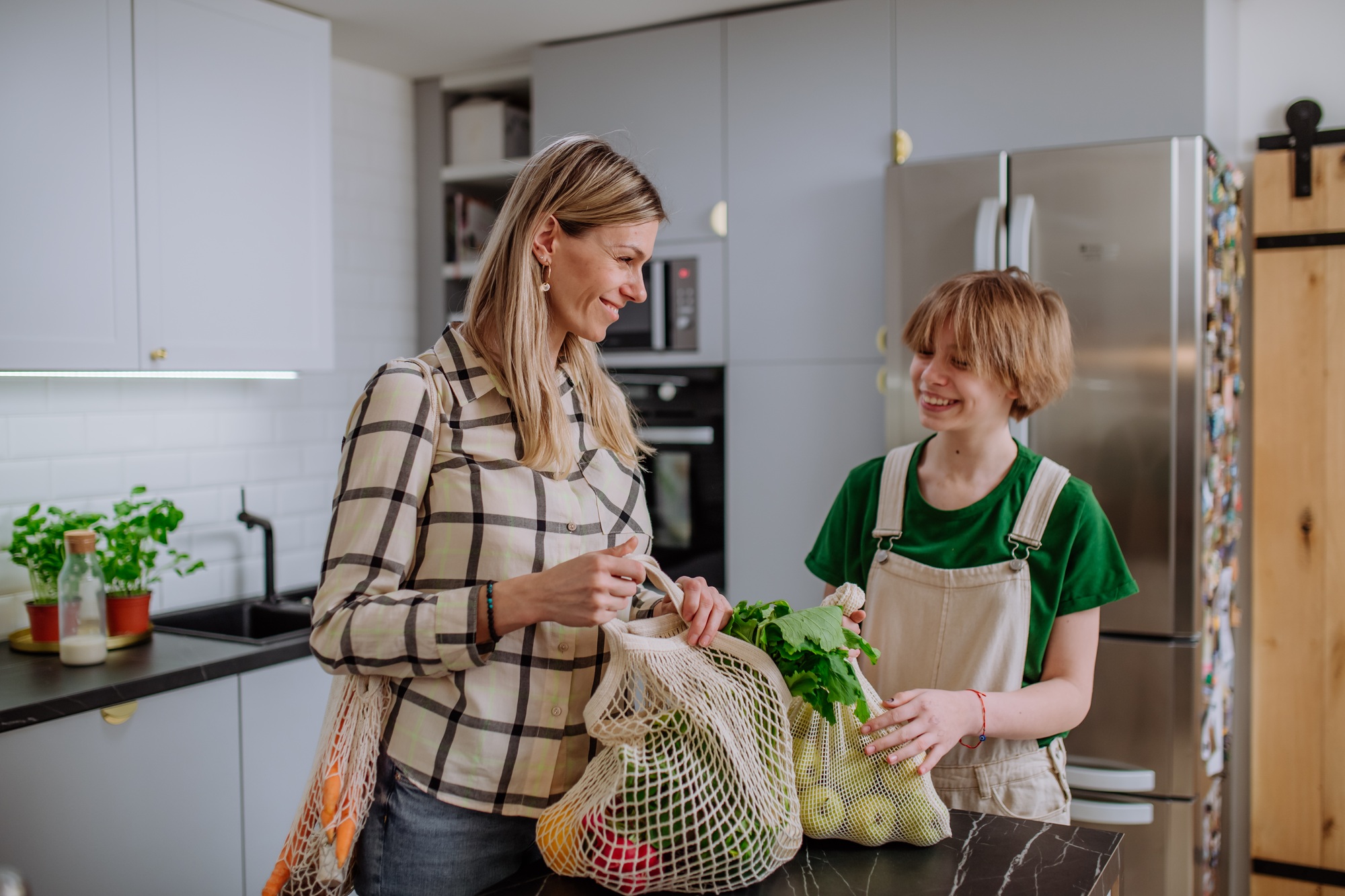 Mother unpacking local food in zero waste packaging from bag with help of daughter in kitchen at