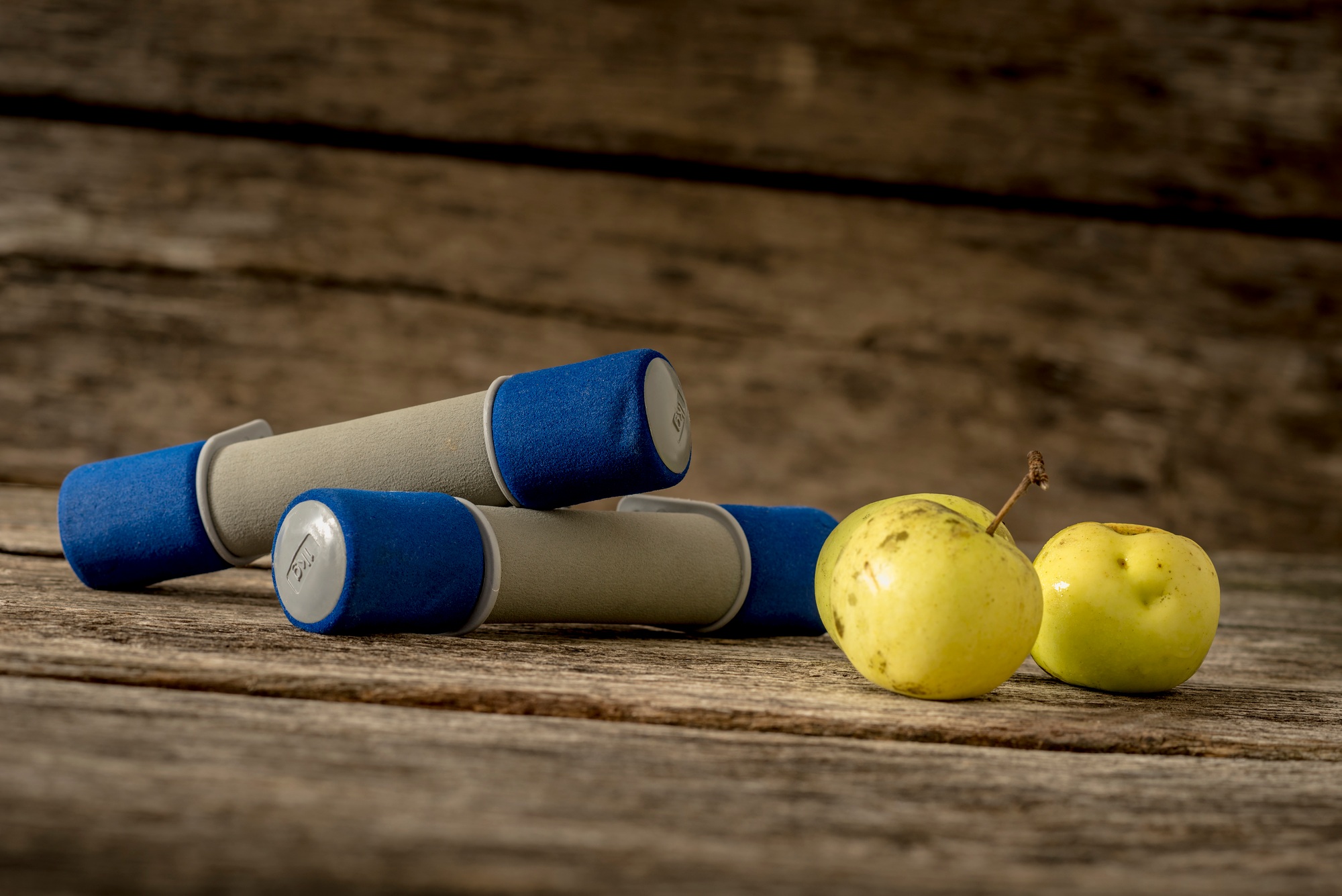 Two workout dumbbells and three fresh ripe apples lying on a textured rustic wooden desk