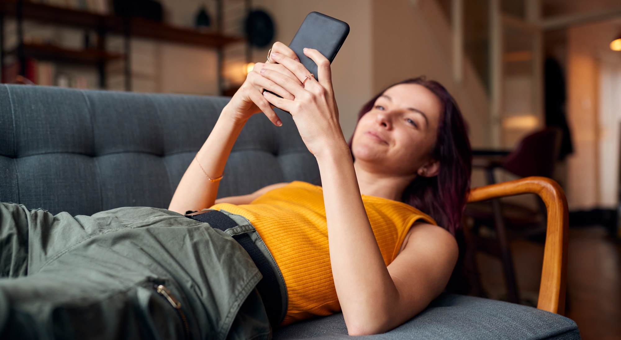 Young Woman Lying On Sofa At Home Looking At Mobile Phone Messages And Social Media