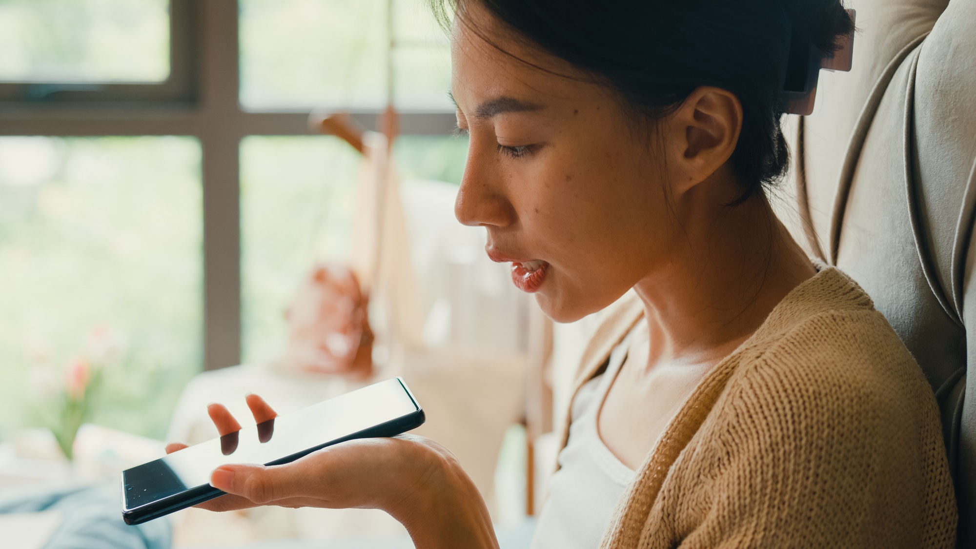 Asian girl with laptop on bed use smartphone giving voice commands to virtual assistant.