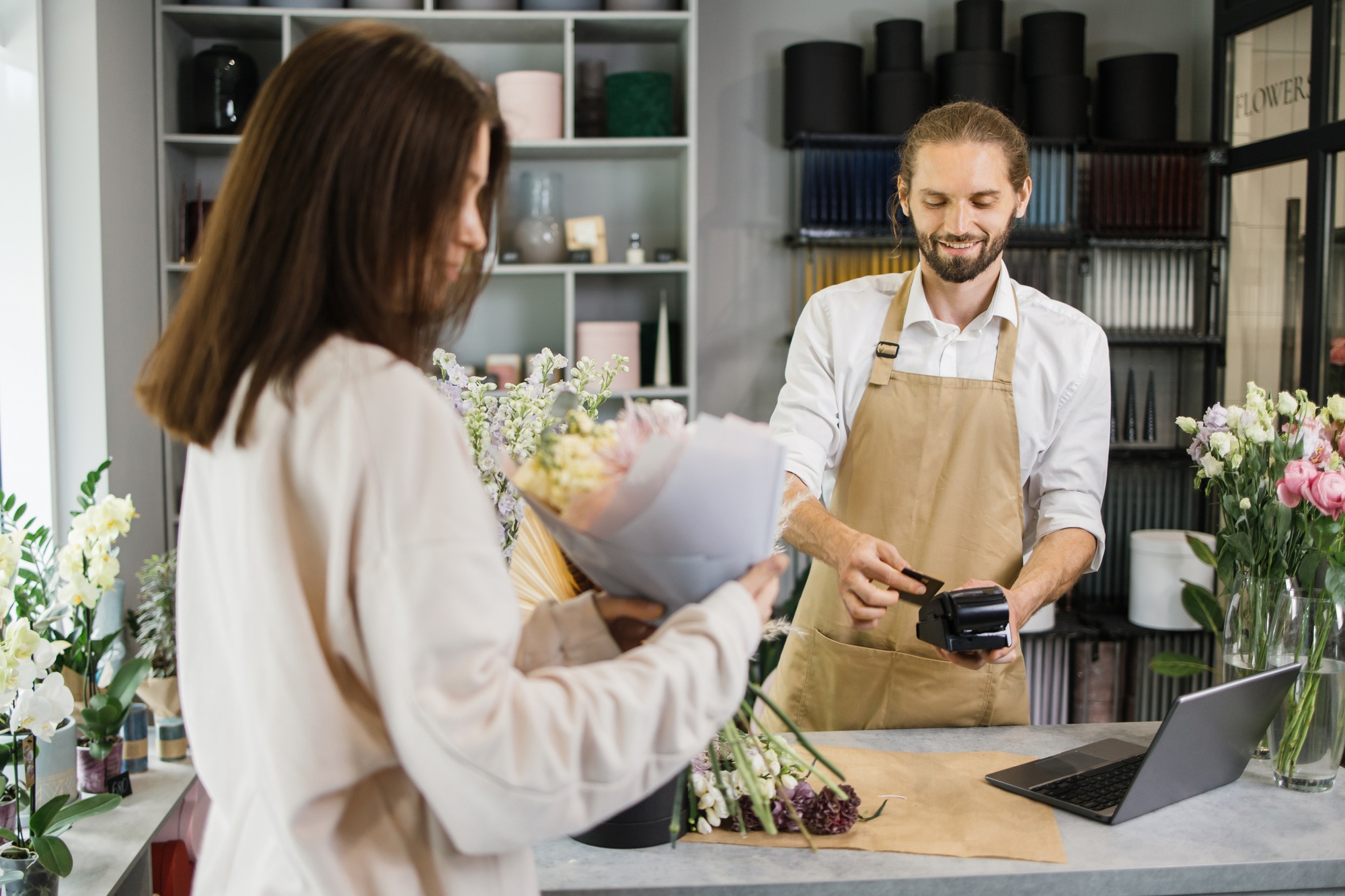 Bearded florist man holding card reader machine at counter with customer paying with credit card.