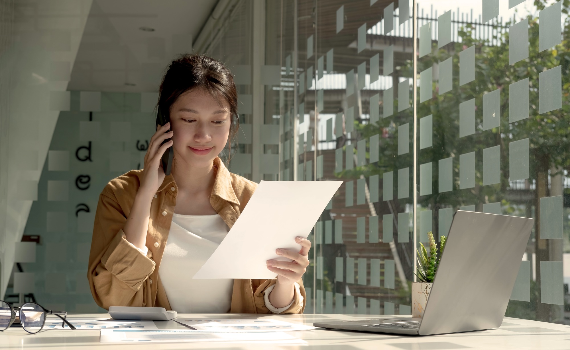 Charming Asian woman with a smile standing holding papers and mobile phone at the office.