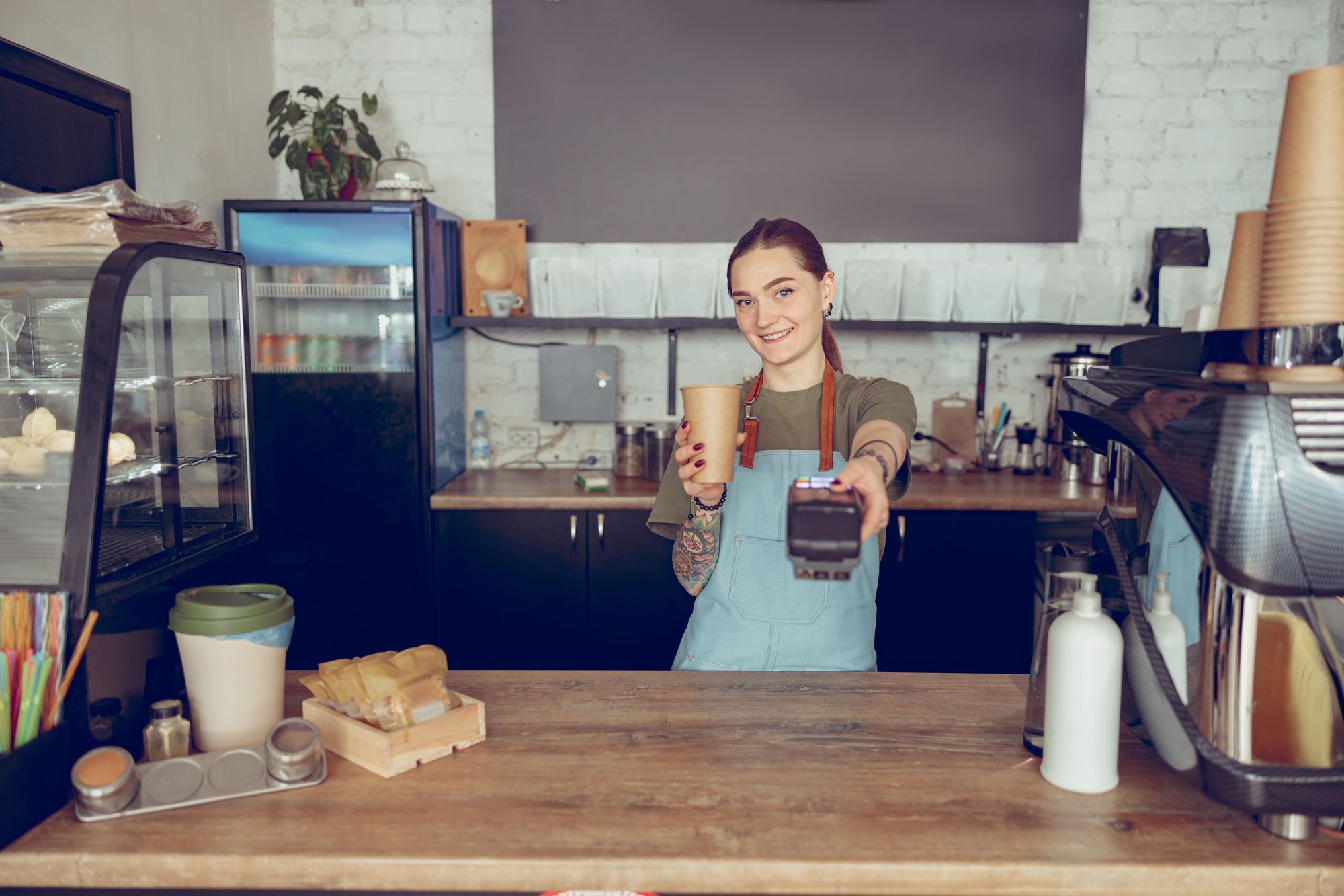 Cheerful woman barista holding contactless card reader and coffee cup