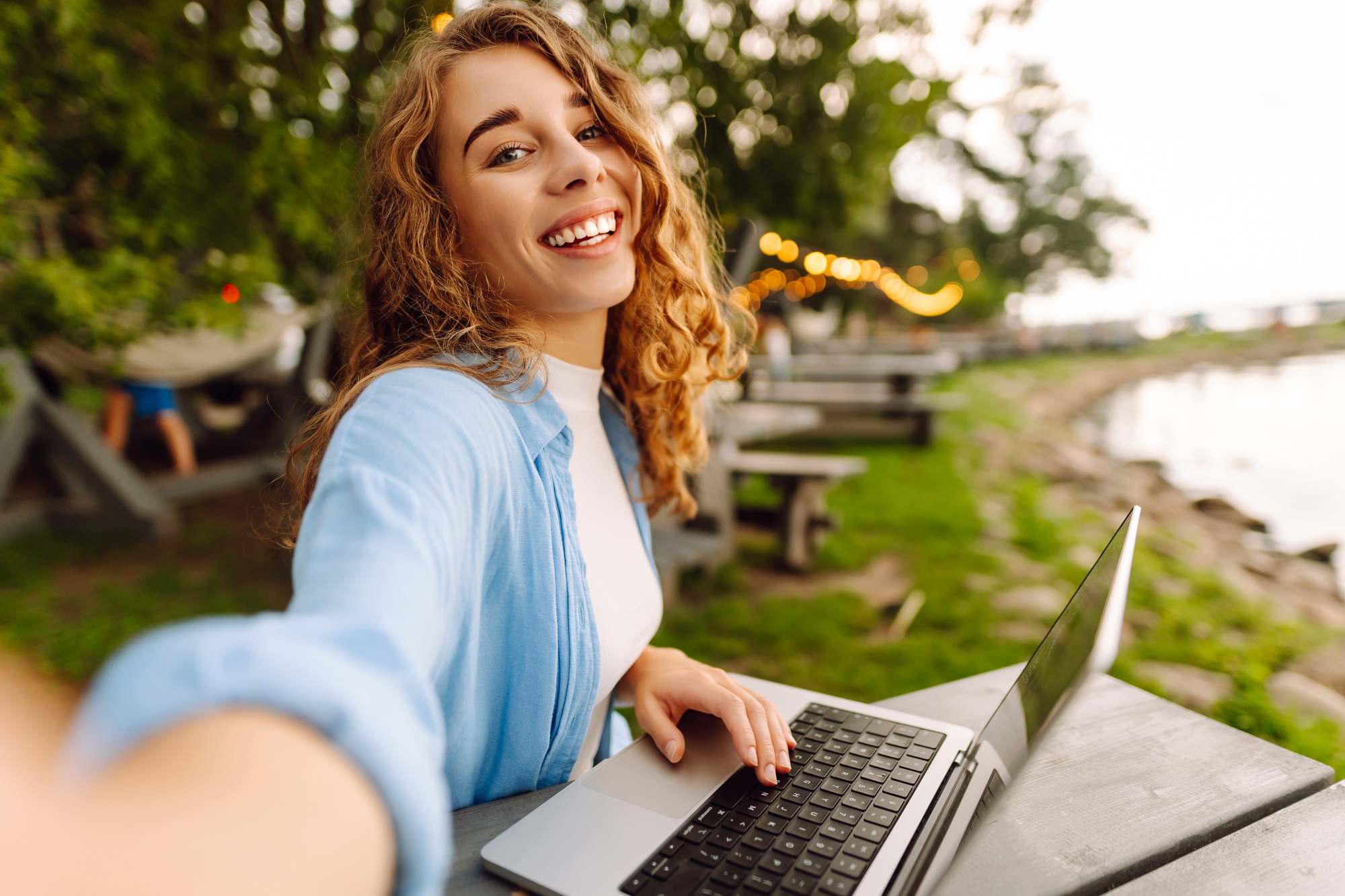 Happy woman in stylish clothes sitting at a table outdoors taking a selfie, working on a laptop.