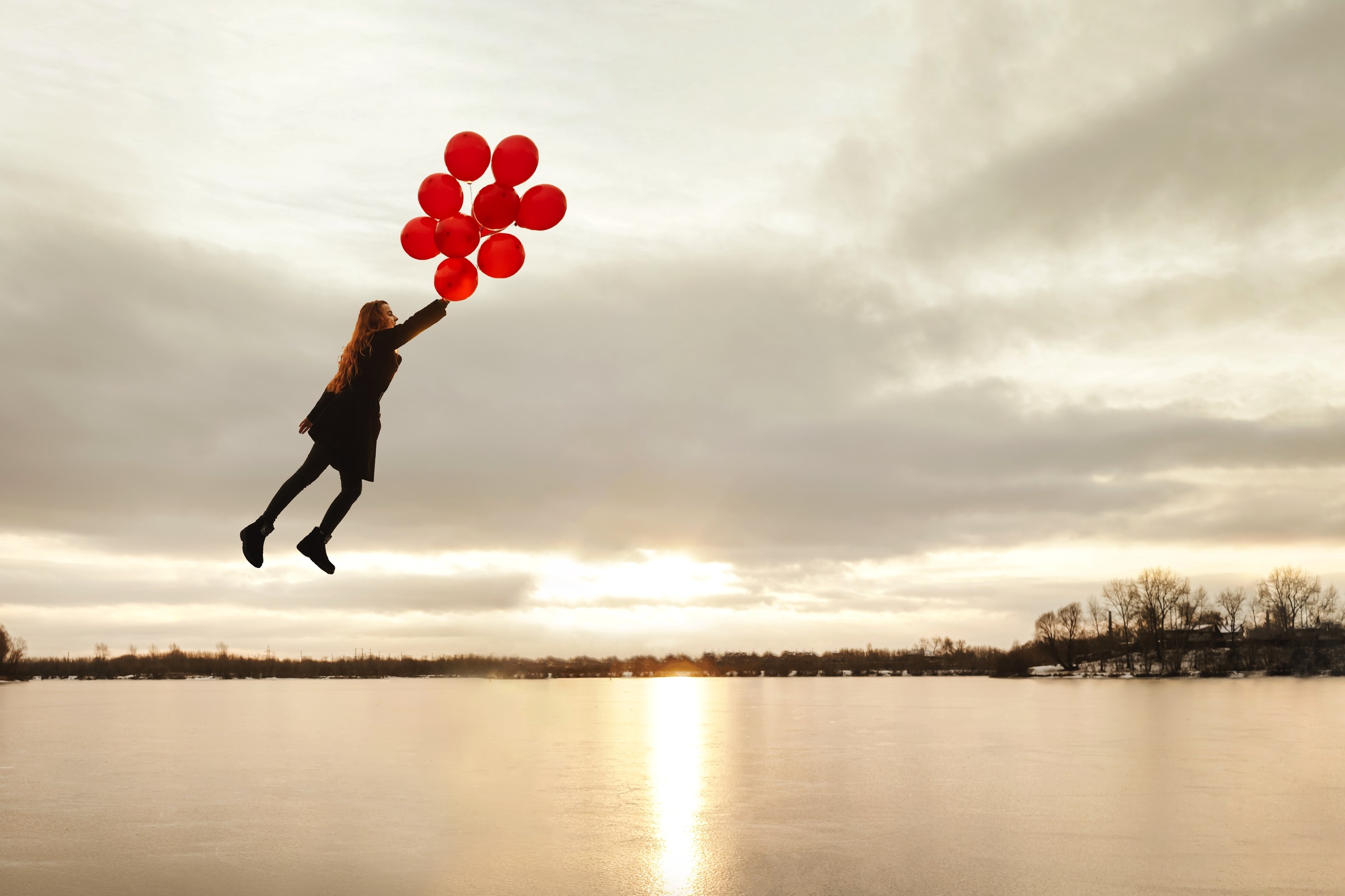 Imagination, happy girl fly with red balloons at sunset on the frozen river, follow your dream.