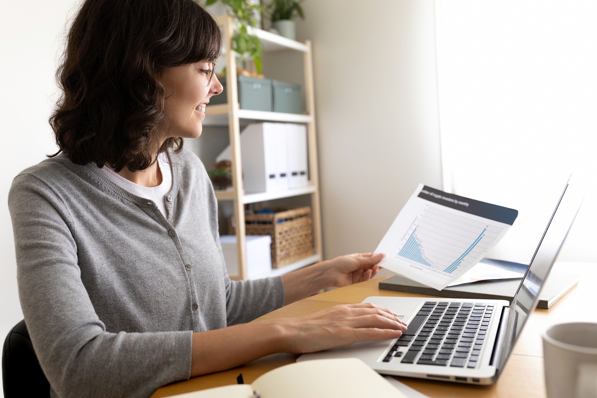 Side view of happy woman looking at documents while working with laptop at home office. Copy space.