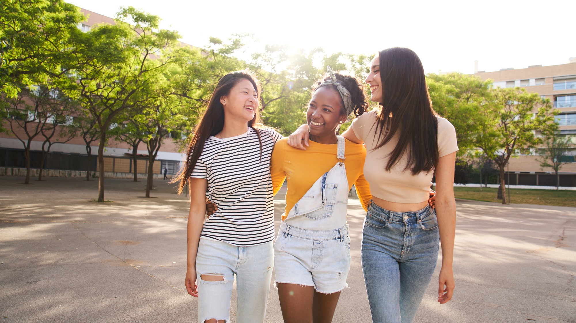 Three cheerful stylish and friendly multi-ethnic girls walking embracing city park laughing together