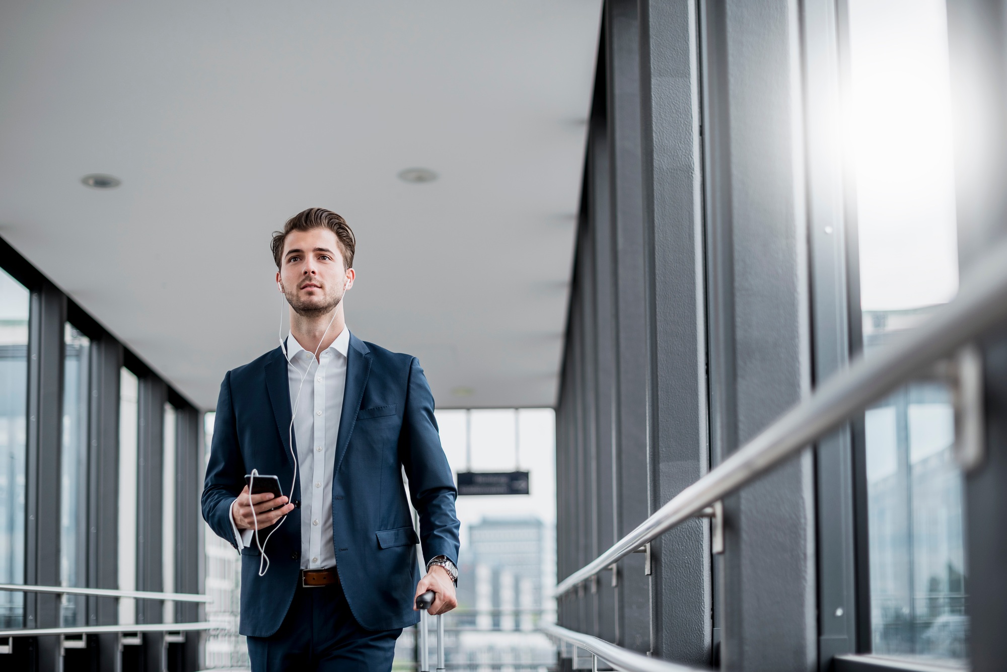 Young businessman in a passageway with cell phone, earbuds and rolling suitcase on the go