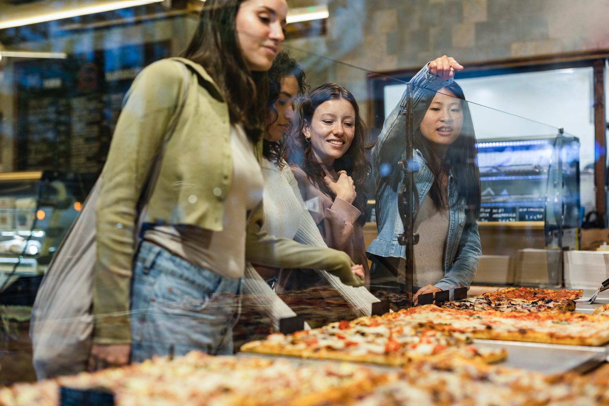 Young women choosing pizza slices at pizzeria display window