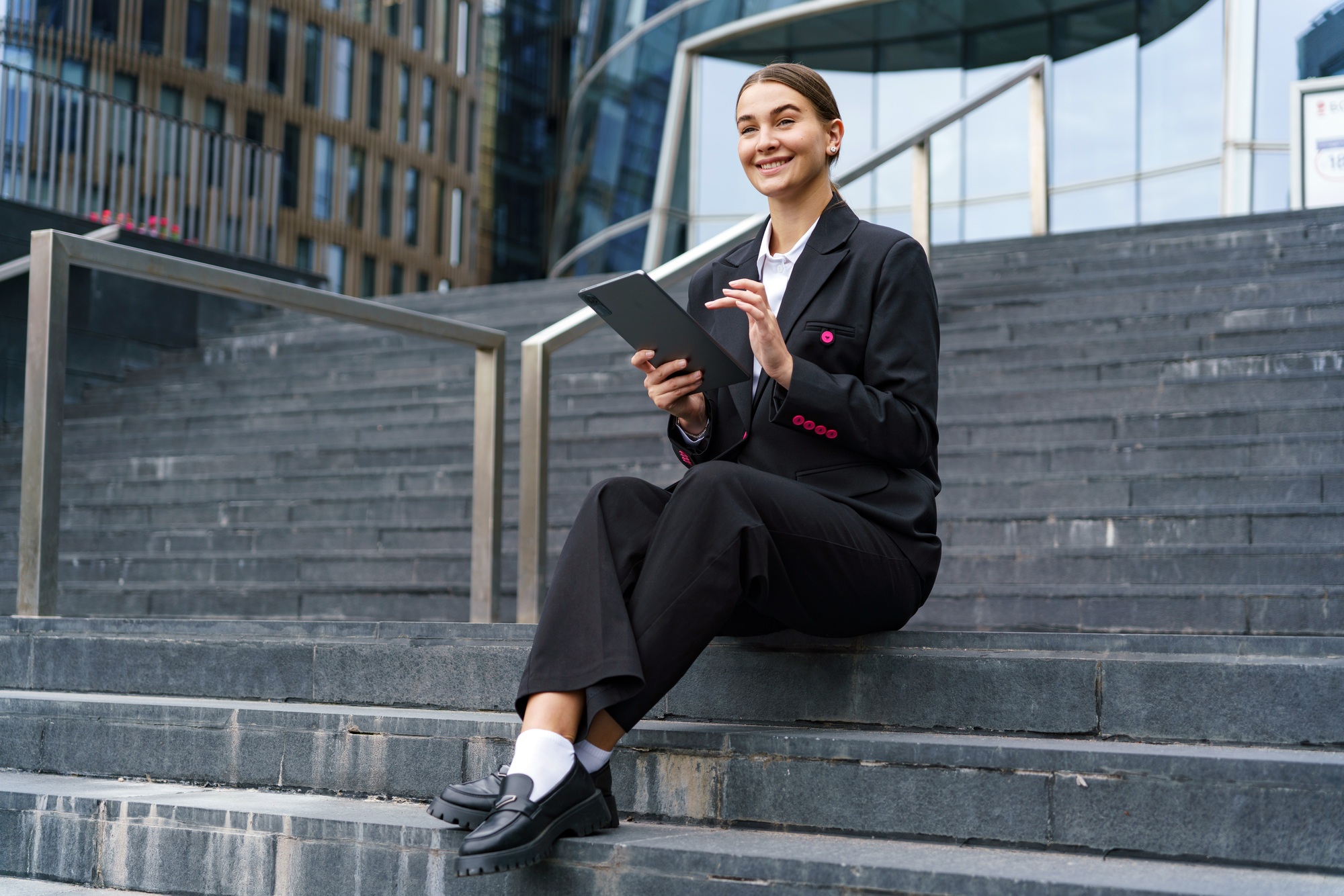 A confident woman in a black suit sits on urban steps, engaging with a tablet, exuding professionali