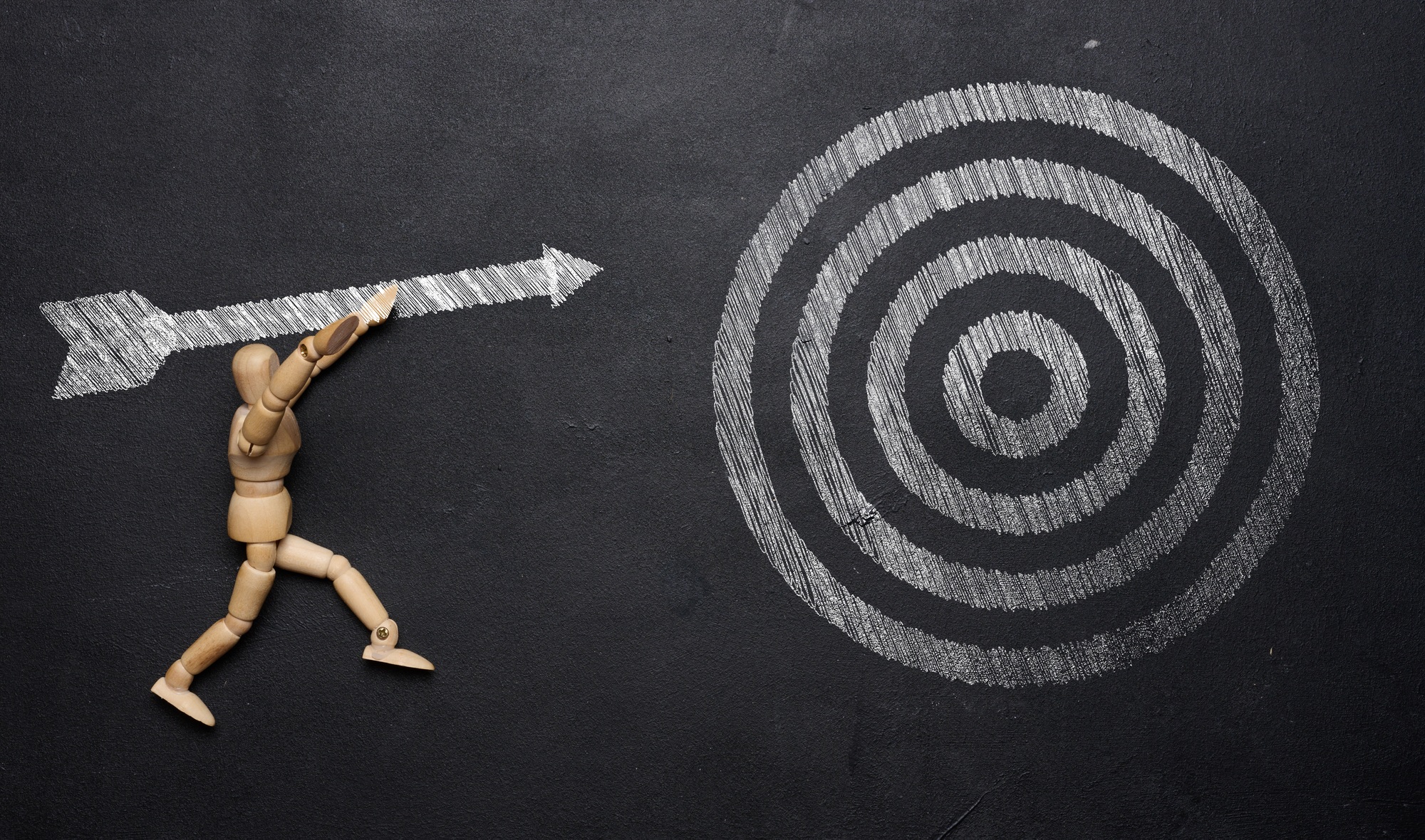 A wooden mannequin holds a drawn arrow and aims precisely at the target on a black chalkboard.