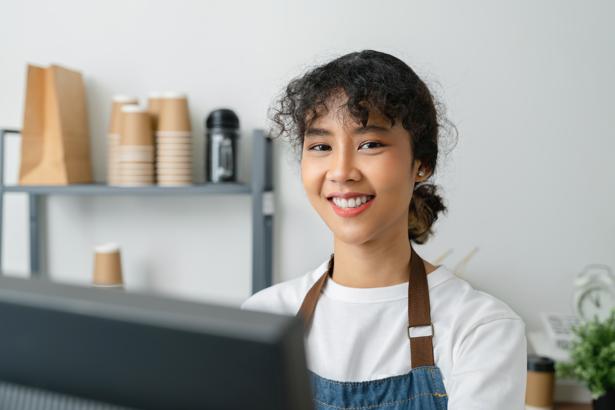 Asian woman cashier wears an apron and using pos terminal to input orders on coffee shop counter.