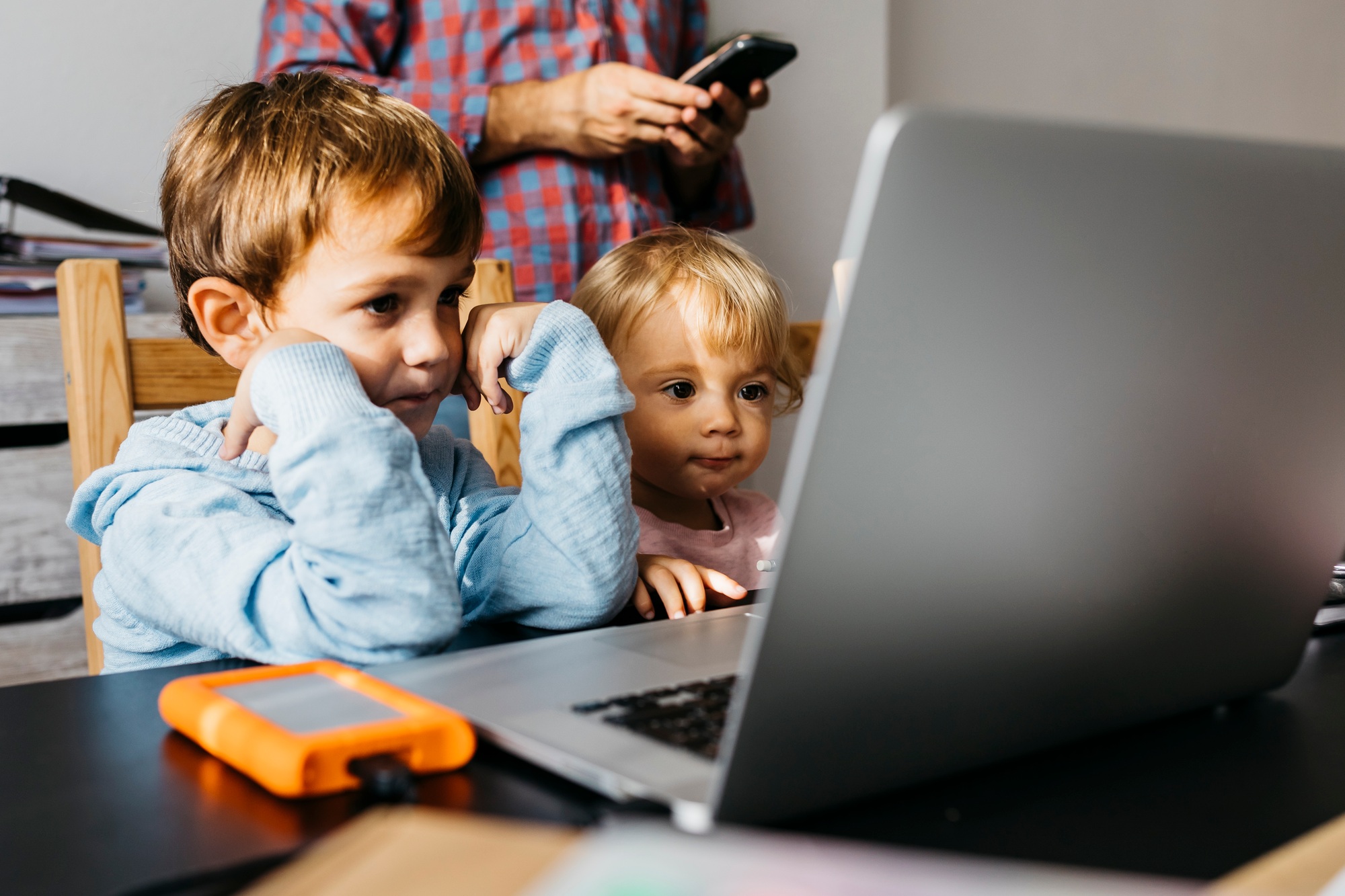 Brother and sister watching a video on a laptop, father using smartphone in background