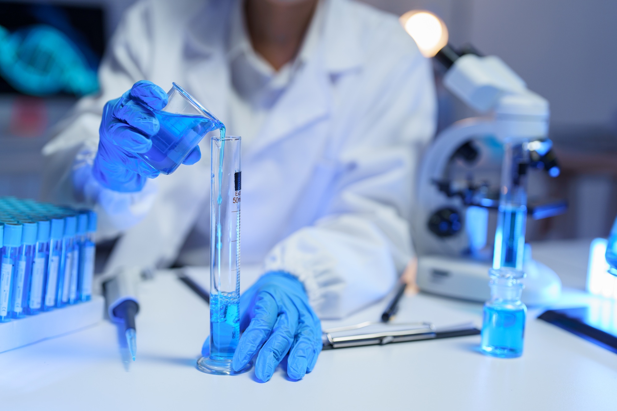 Close-up of a scientist in gloves pouring vibrant blue liquid from a beaker into a graduated cylinde