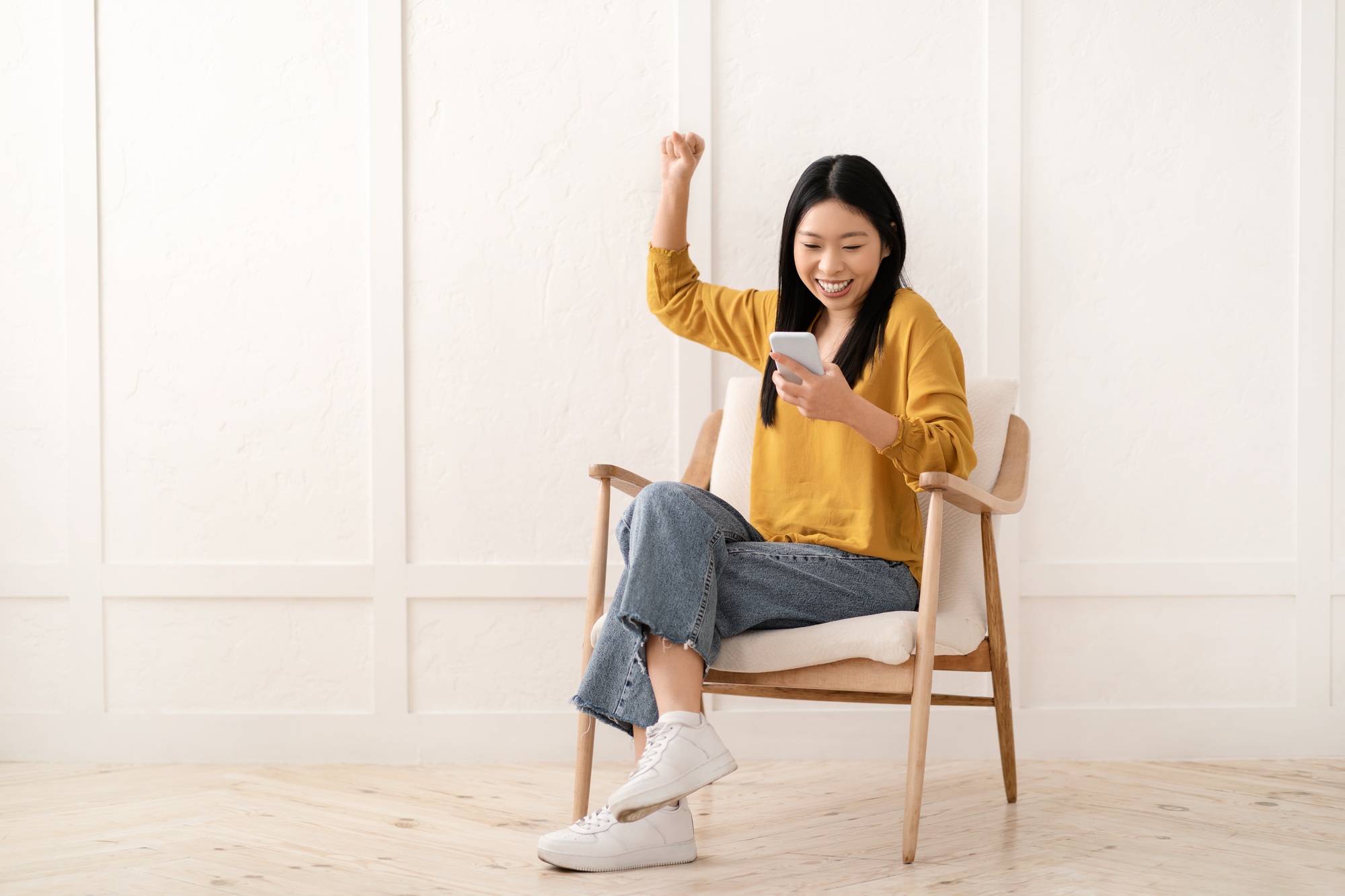 Emotional asian woman sitting in armchair, using cell phone, gesturing