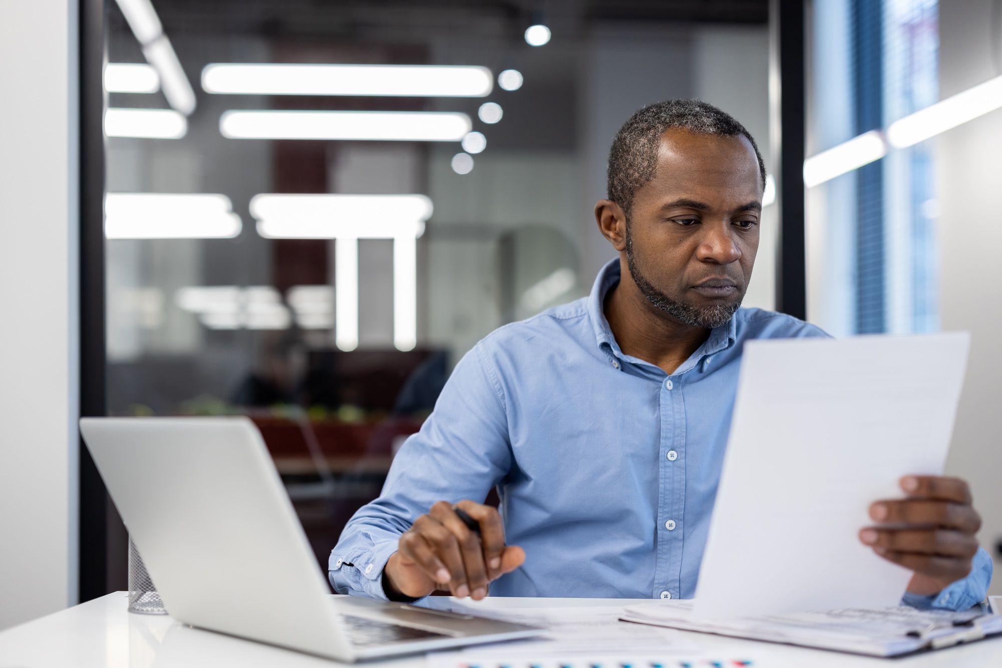 Focused businessman reviewing documents at desk with laptop in modern office environment.