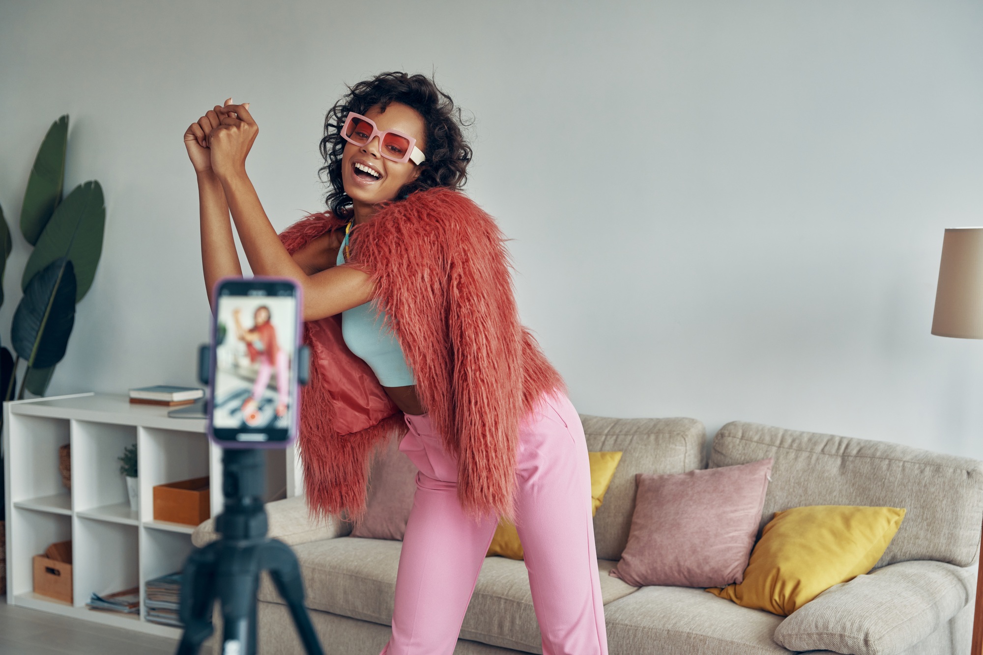 Happy African woman in fluffy jacket creating her dancing video on phone camera at home