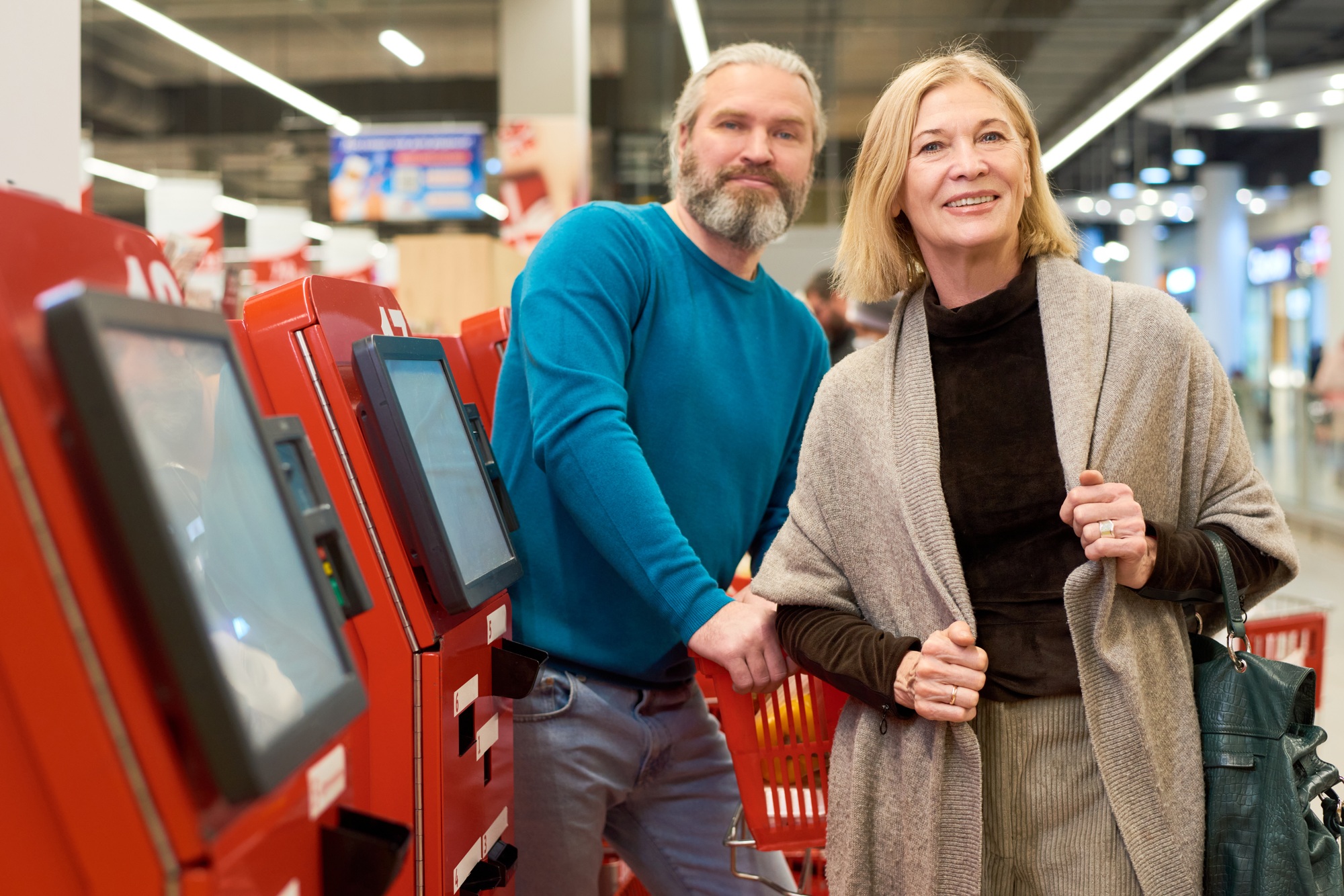 Happy aged couple in smart casualwear standing by self service terminals