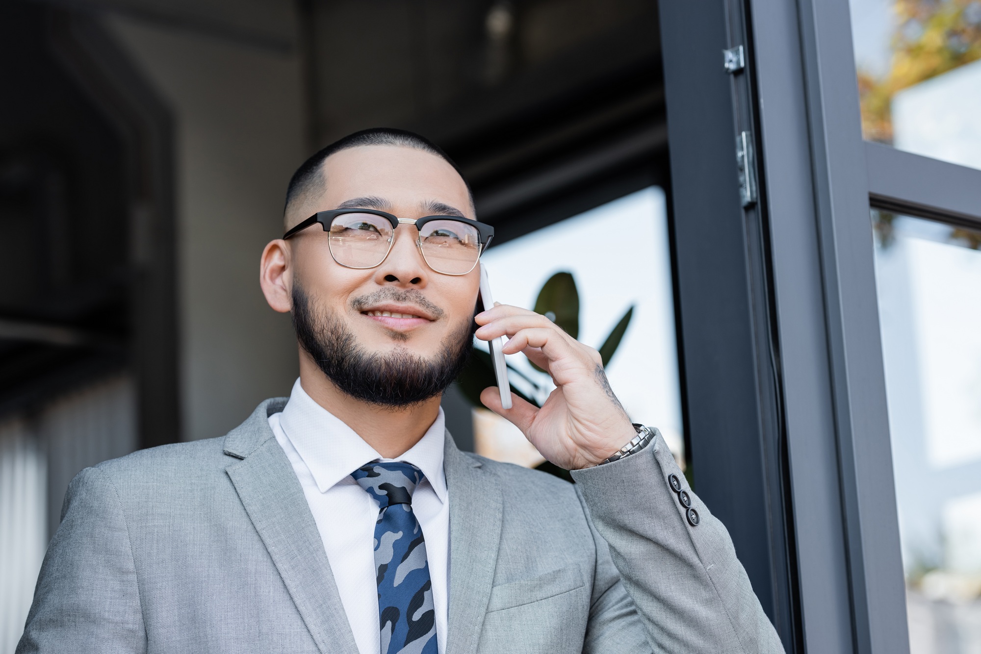 happy asian businessman in eyeglasses talking on mobile phone near window