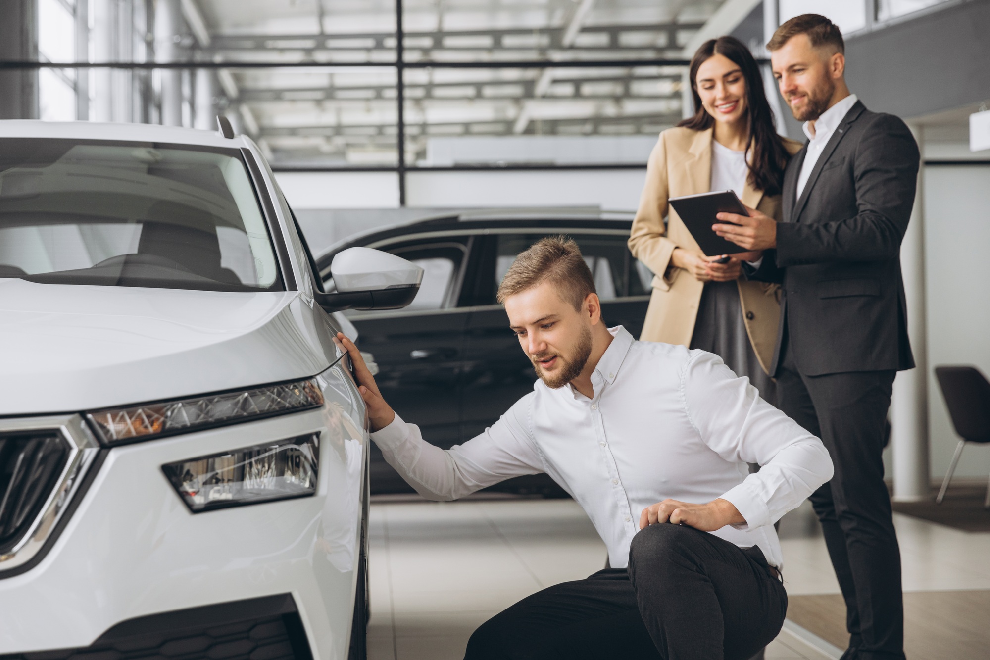 Happy modern man inspecting new car in ?ar showroom and in background the manager of car showroom