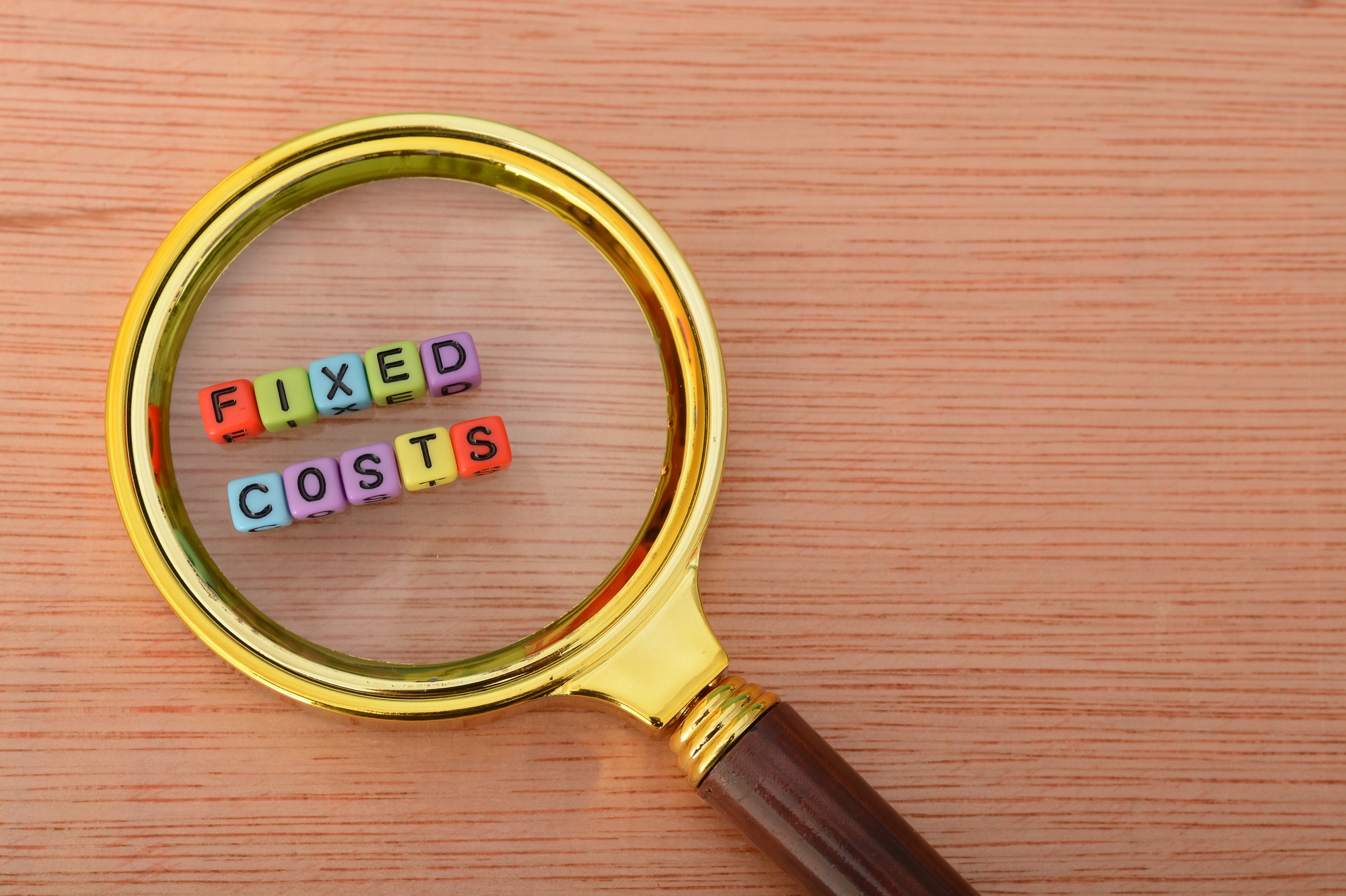 Magnifying glass with text FIXED COSTS.. Examples of fixed costs include rent, salaries, insurance,