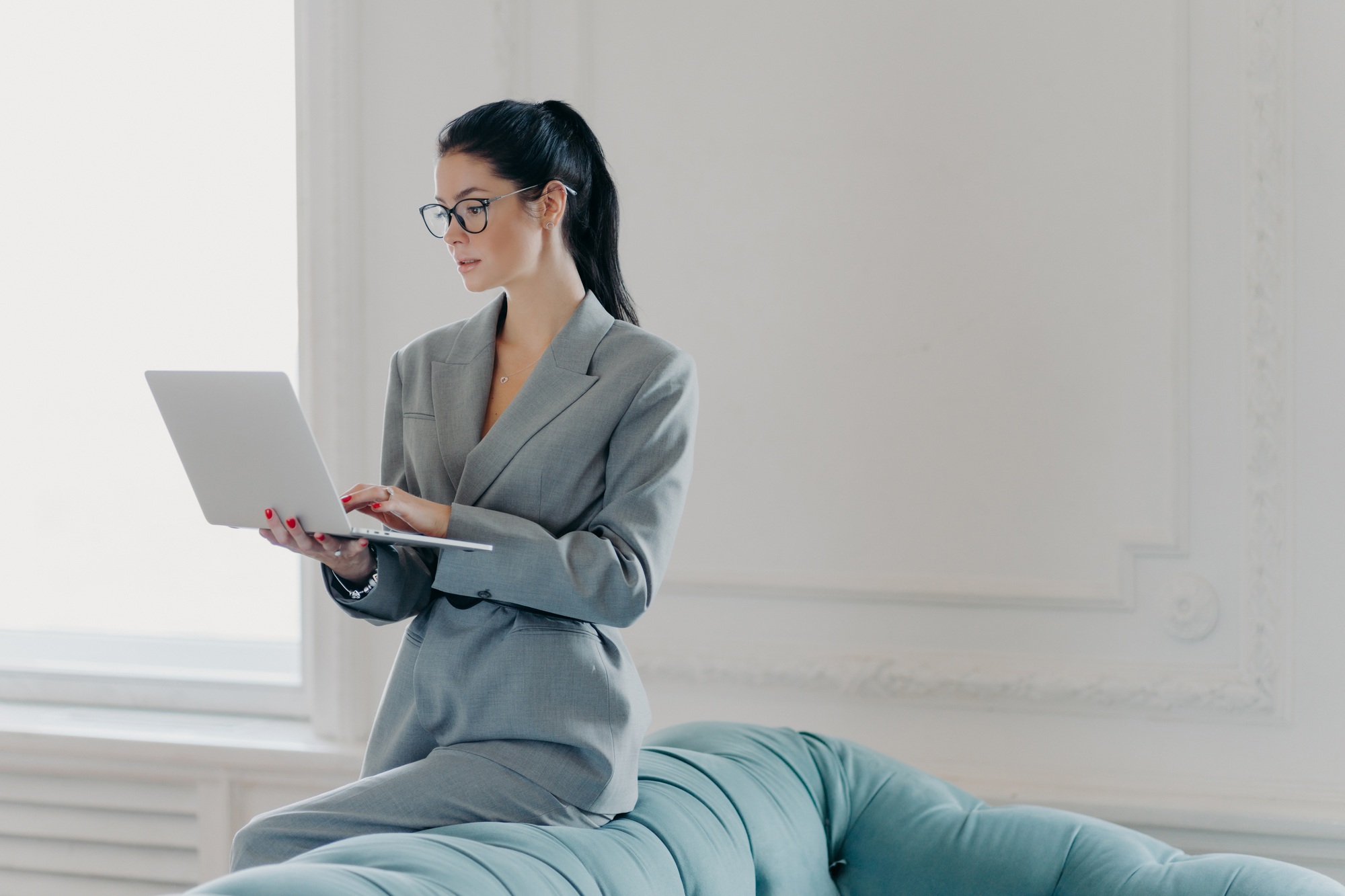 Photo of brunette executive worker poses in home office near sofa, holds laptop computer