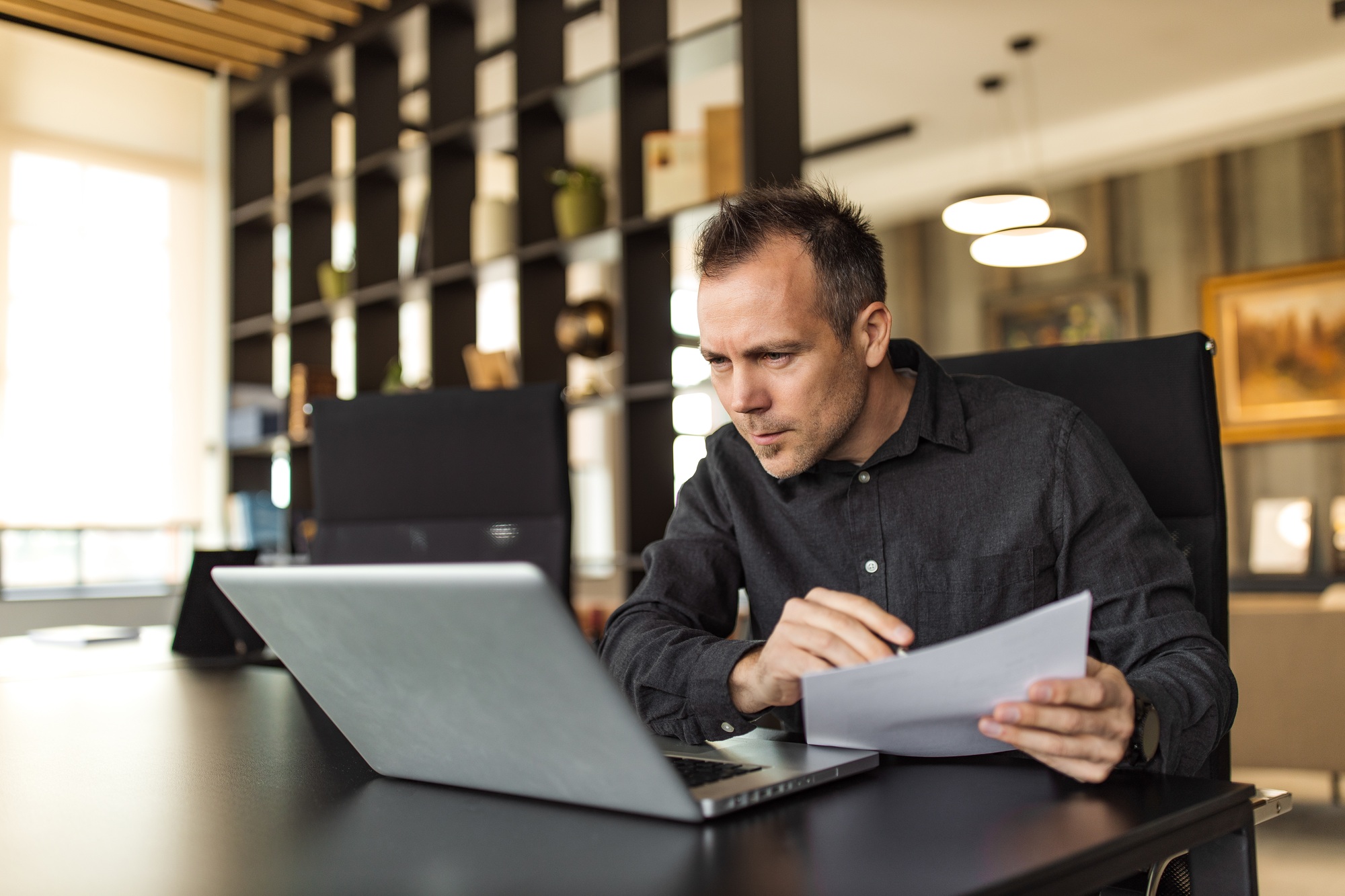 Portrait of a serious businessman making comparison, holding paper and looking at laptop.