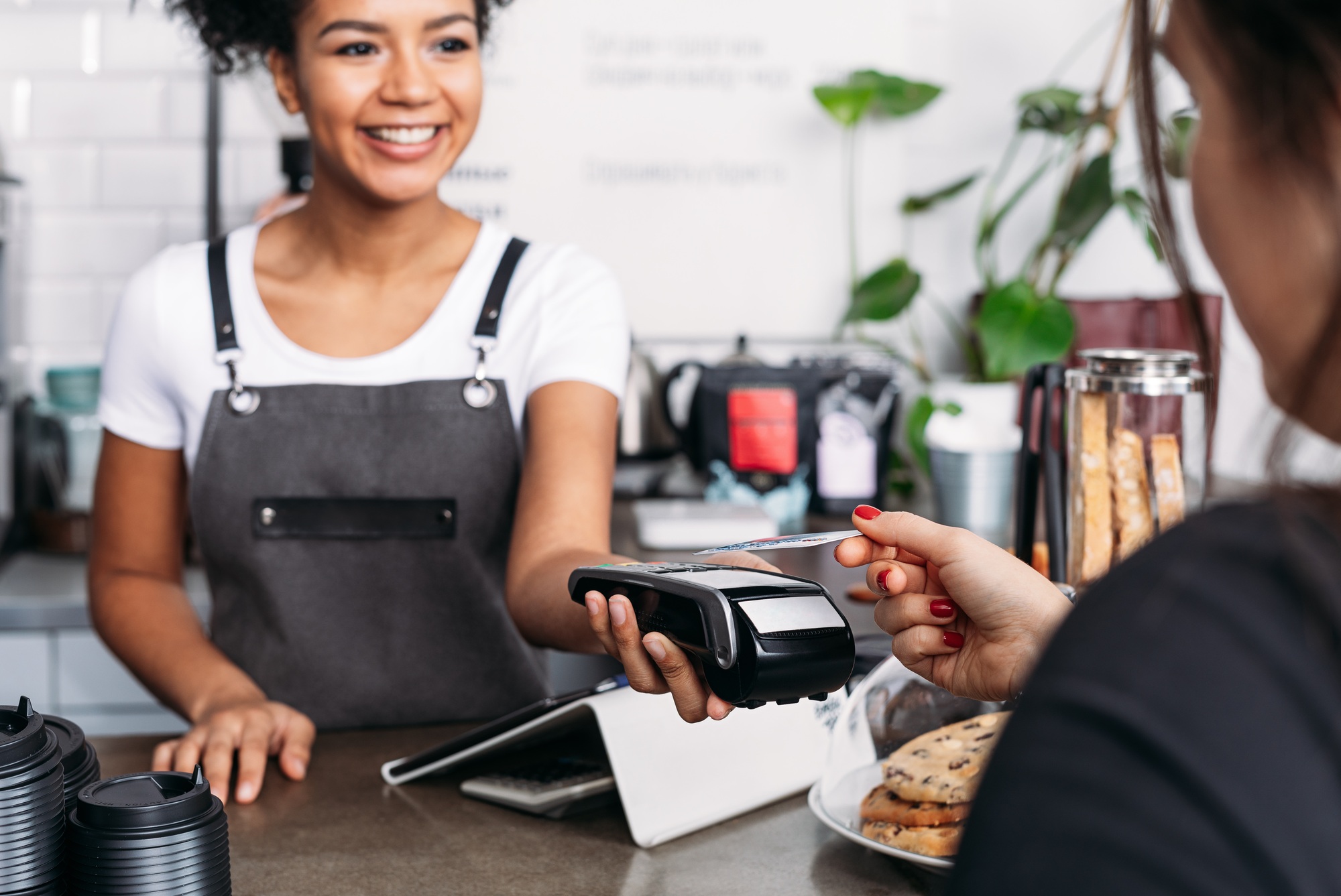 Smiling barista in apron holding POS terminal while customer paying by card