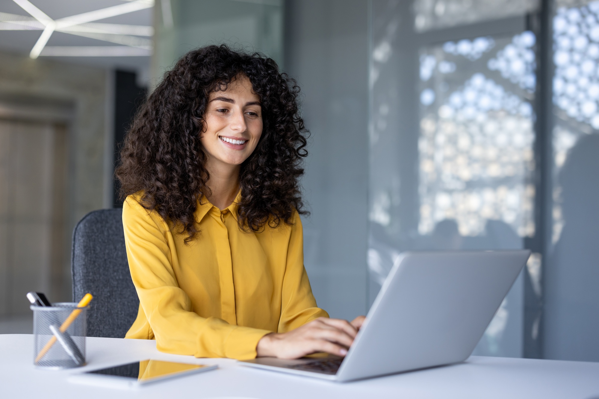 Smiling Hispanic businesswoman working on laptop in modern office, showcasing leadership and