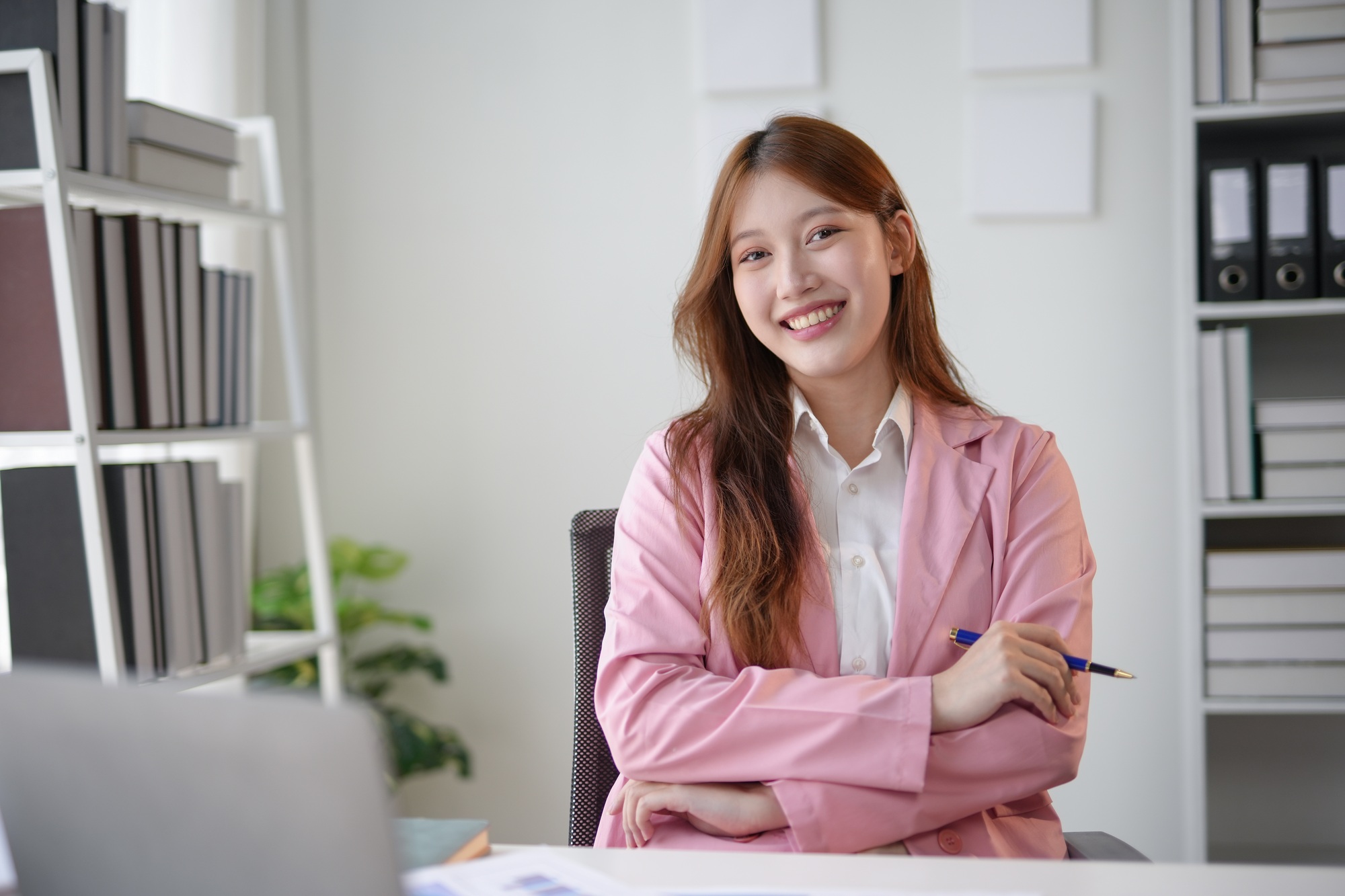 Smiling woman in pink blazer sitting at an office desk with arms crossed