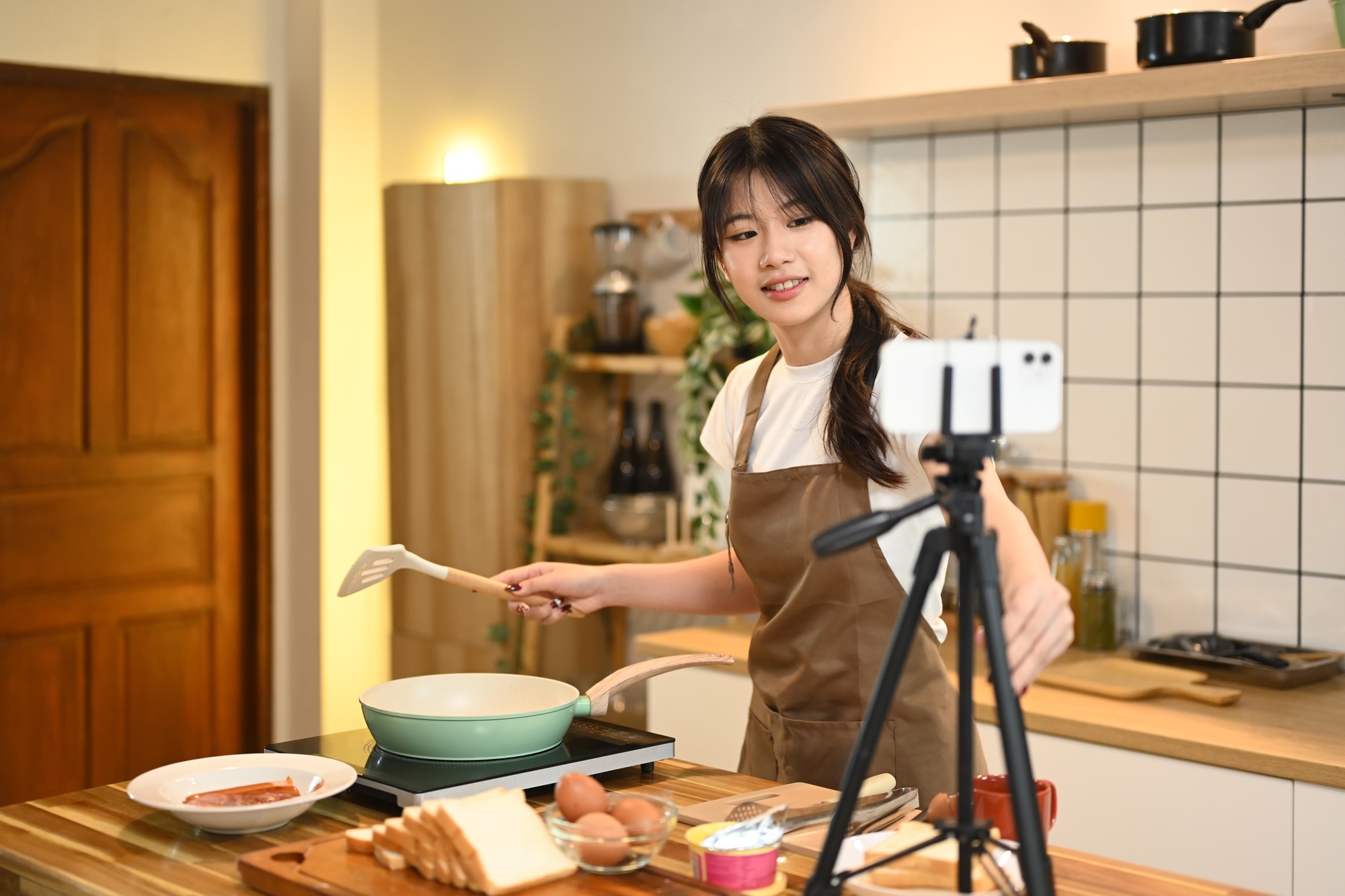 Smiling young woman holding spatula, recording cooking video in her home kitchen