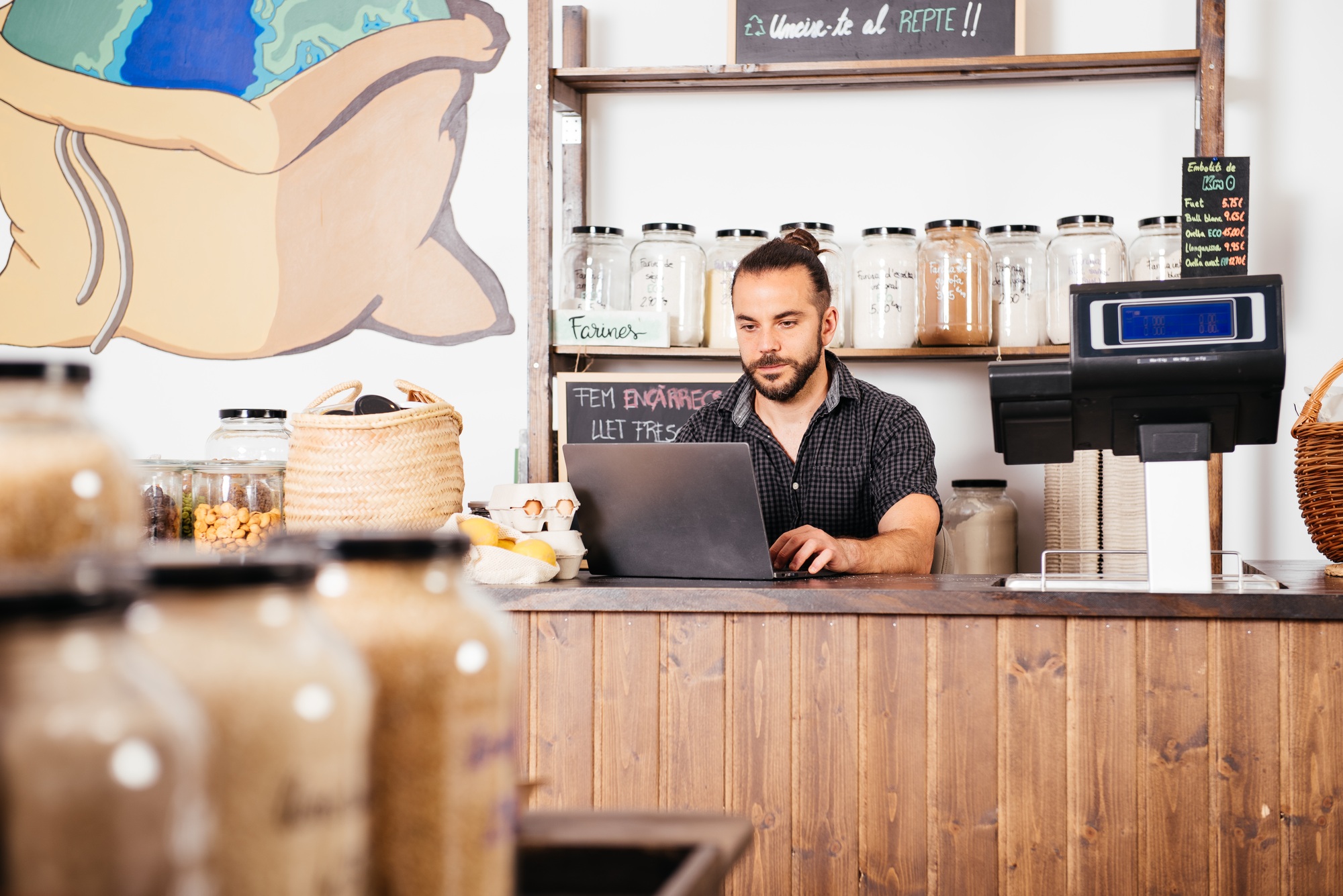 Storekeeper Manages Inventory on Laptop Behind Grocery Counter in the Afternoon