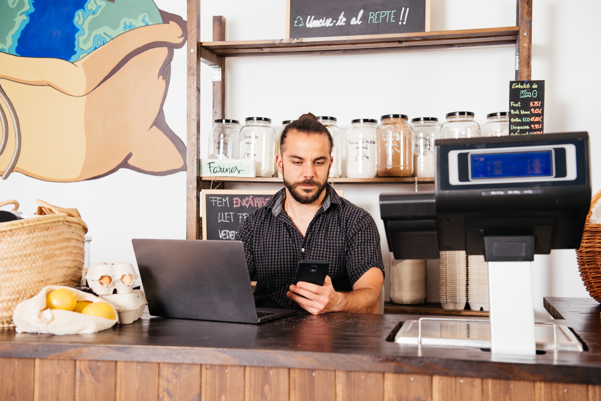 Storekeeper Managing Inventory With Laptop and Smartphone at Grocery Counter During Work Hours