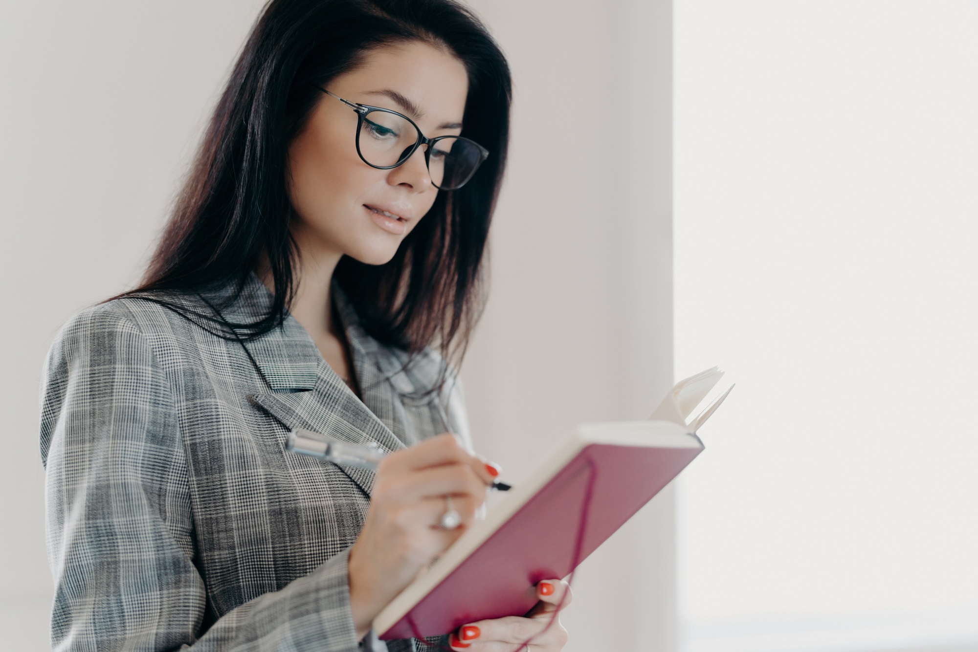 Thoughtful woman with glasses immersed in reading and taking notes in a planner.