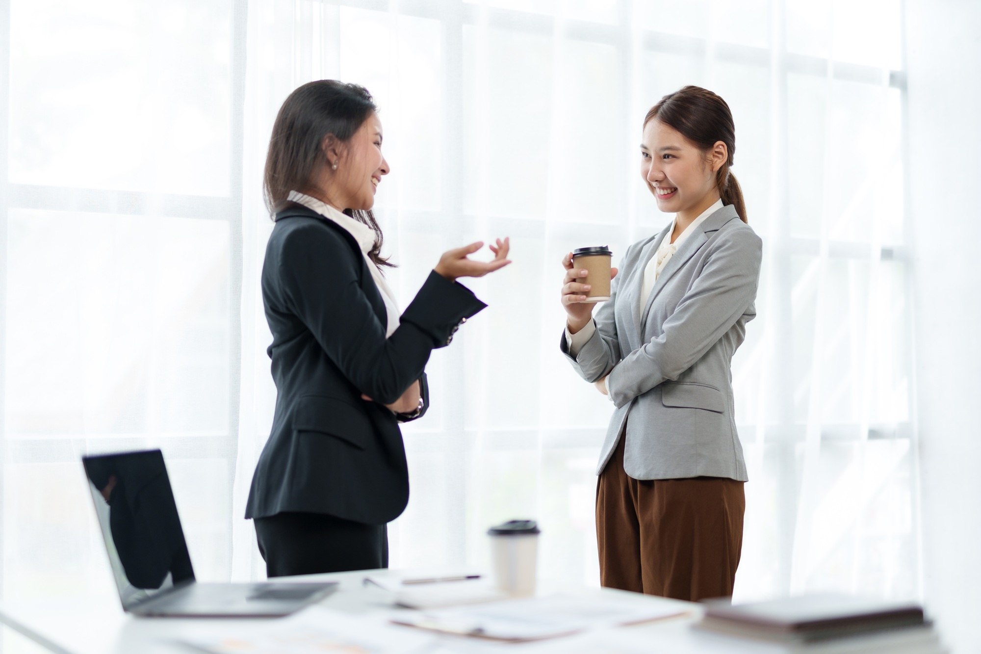 Two beautiful Asian business women standing chatting during a break relaxing coffee, discussing work