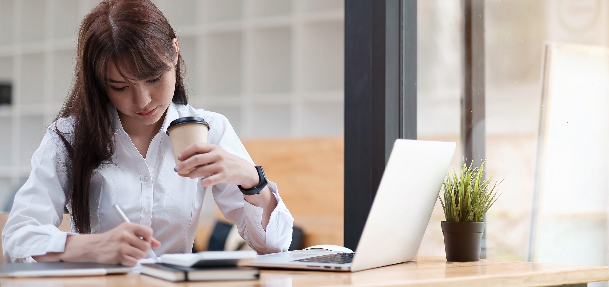 Woman having a tea, sitting on a floor in an office, writing down notes, opened laptop in front of