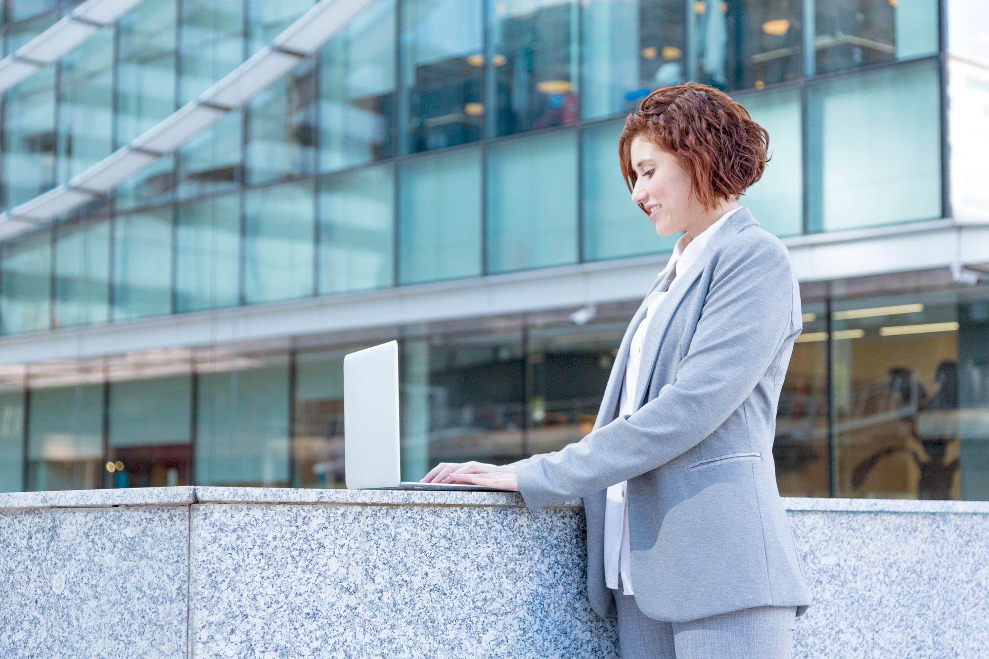 Young executive woman in gray suit working outdoors with laptop