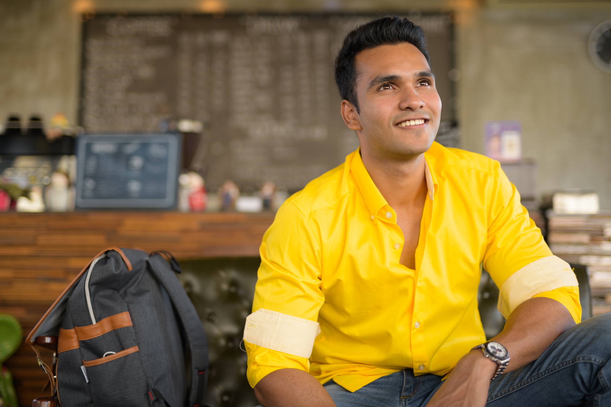 Young happy Indian man thinking and sitting at the coffee shop