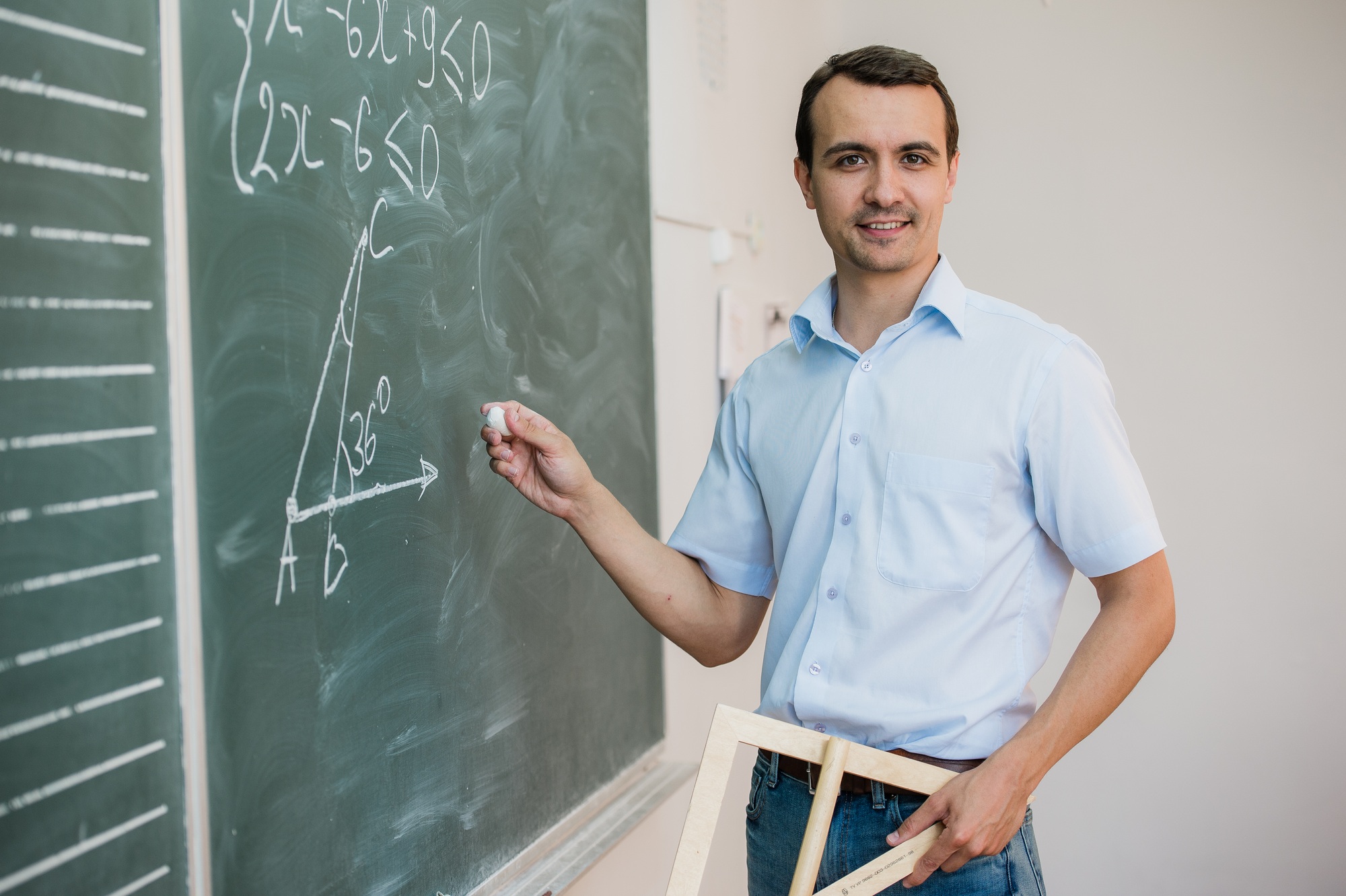 Young teacher or student holding triangle pointing at chalkboard with formula, looking to camera and