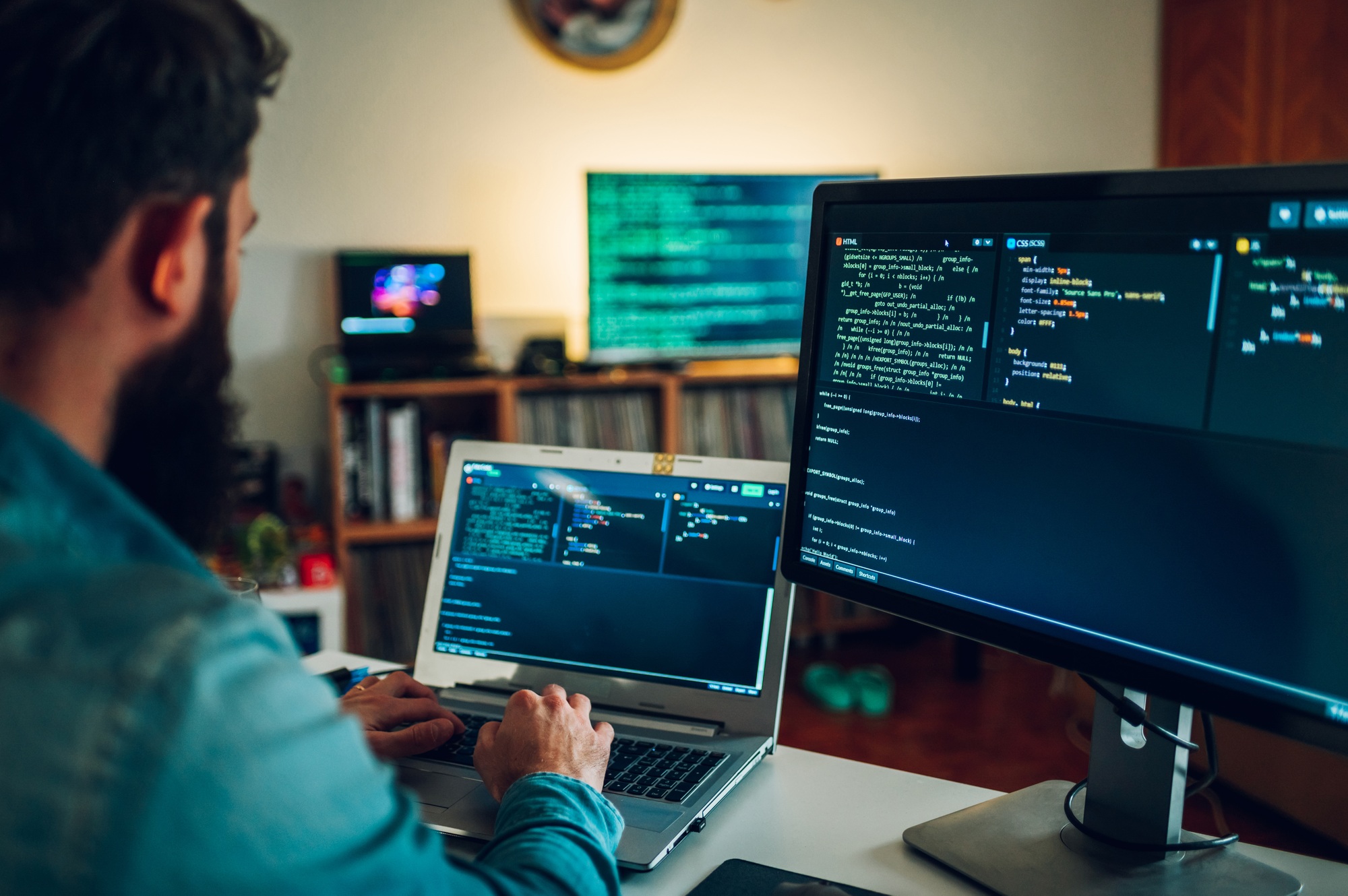 A database scientist is entering data on his laptop while sitting in a home surrounded by screens.
