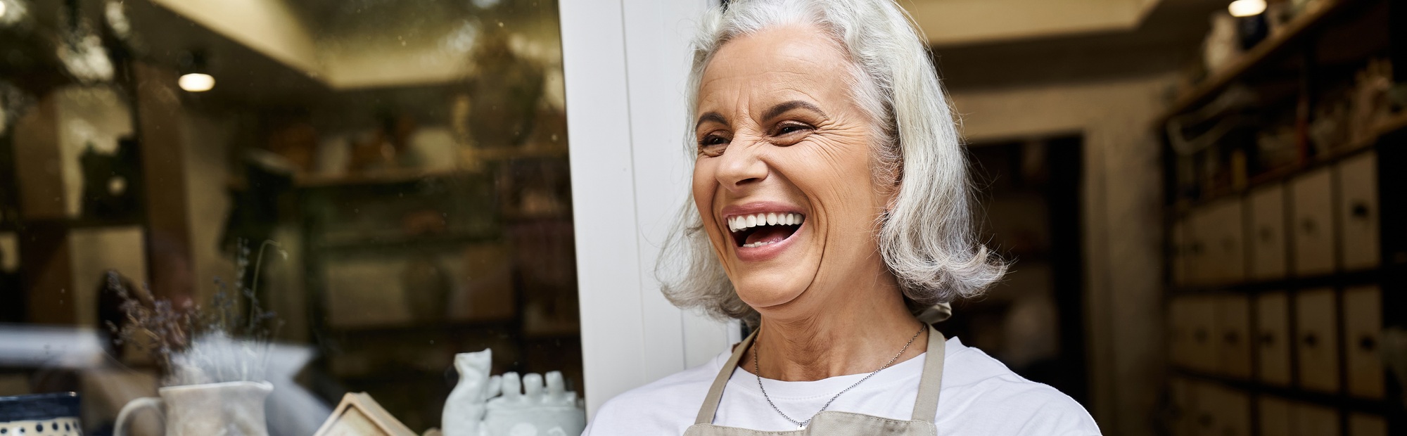 A joyful mature woman smiles warmly while holding a handmade item in a cozy shop