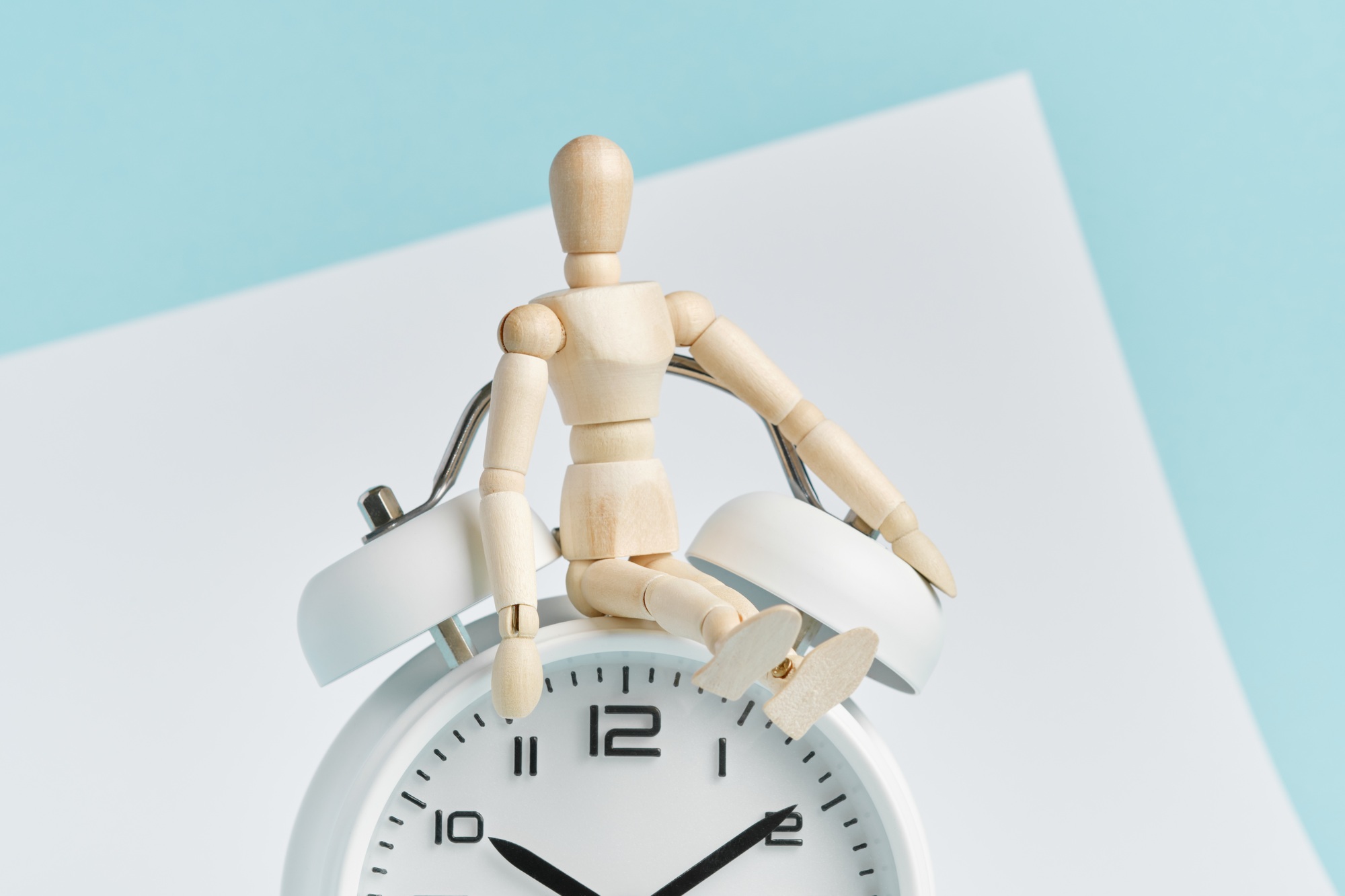 A wooden man sits on a white alarm clock. Time management, time control, punctuality.