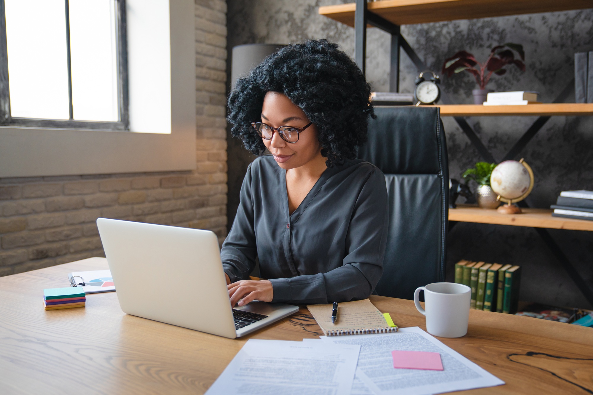African woman in eyeglasses sit at desk works at laptop looks concentrated search solution or ideas