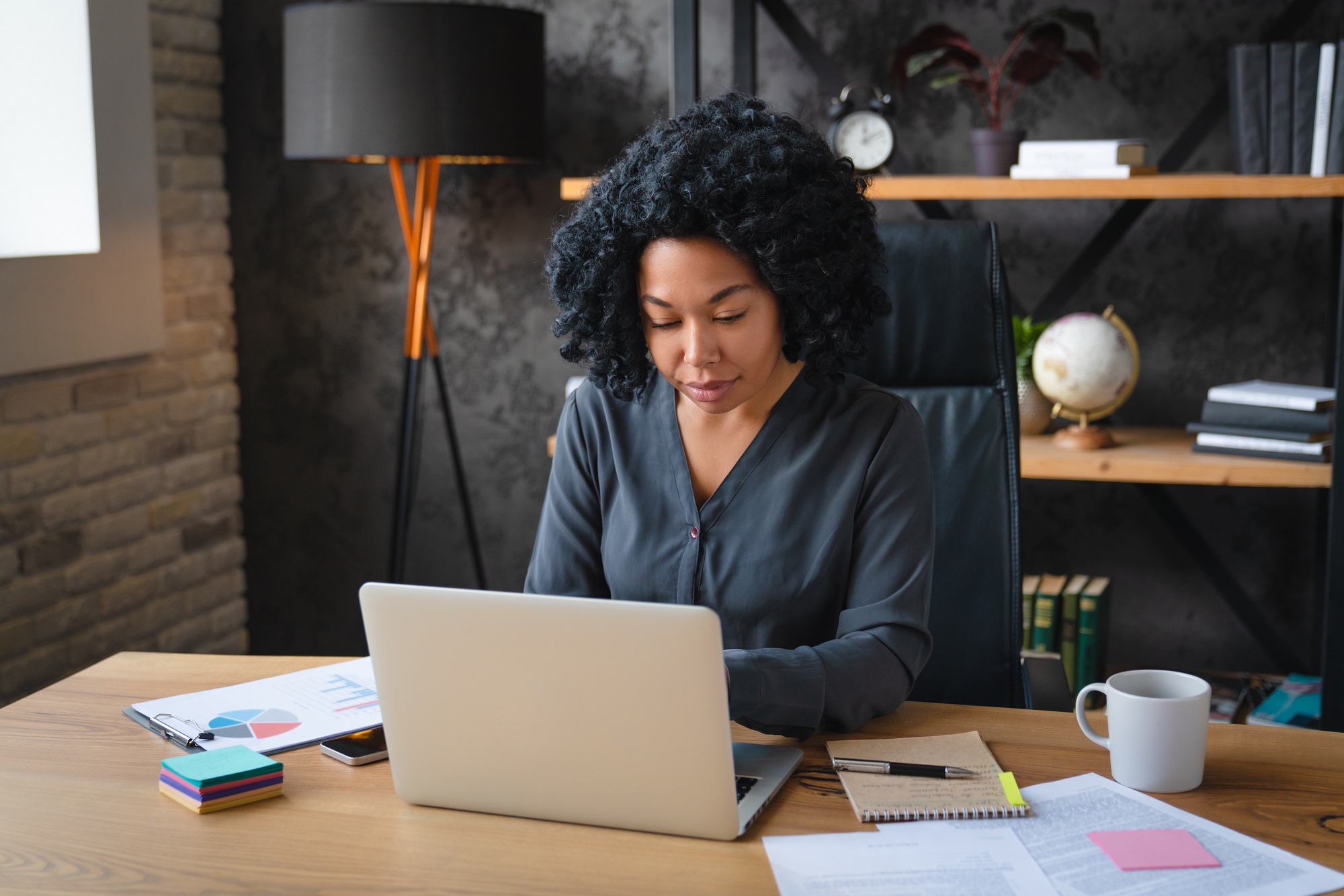 African woman sit at desk staring at laptop screen makes assignment working looks concentrated