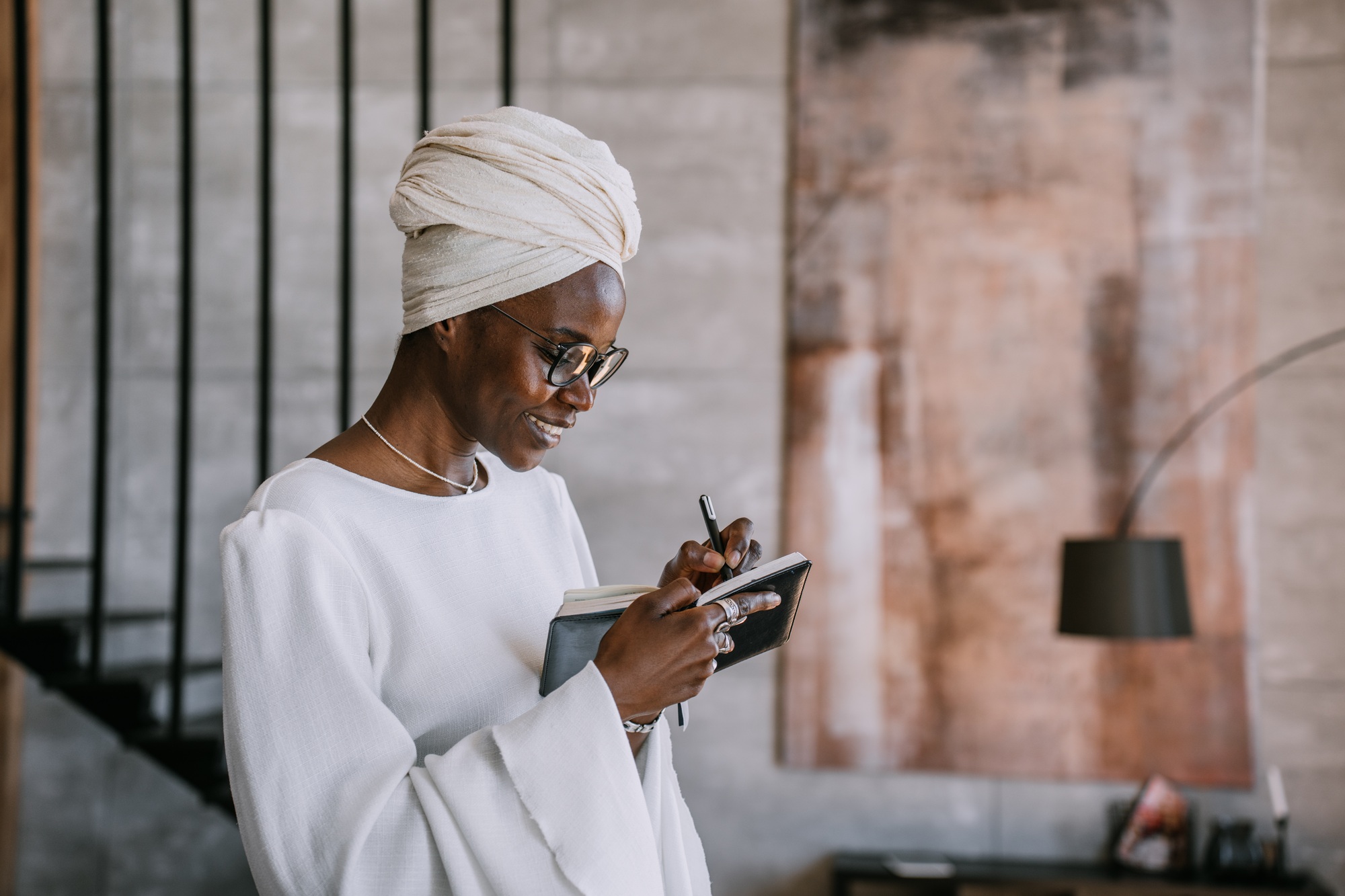 Cheerful African woman in white turban, white dress, glasses holding planner writing notes, smiles.